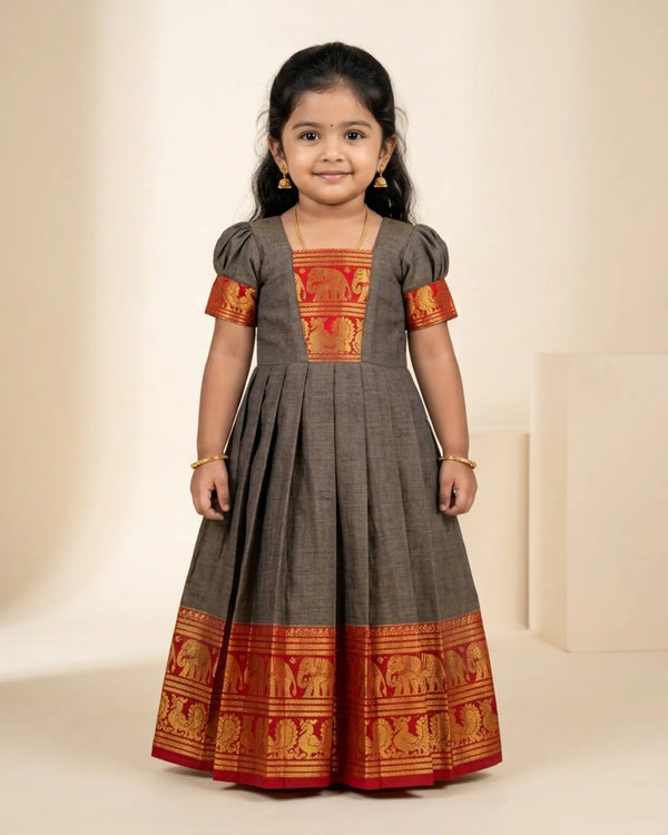 A young girl modeling a dark gray Anarkali gown with red and gold elephant-patterned borders and sleeve cuffs, set against a warm beige studio background.