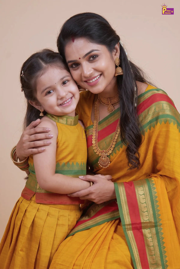 A woman and a young girl wearing matching traditional Indian attire, featuring a mustard yellow saree and dress with green and red borders, posed together against a solid beige background.