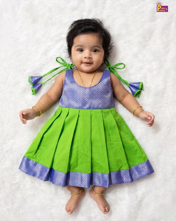 A baby girl lying on a white textured blanket, wearing a sleeveless green and purple dress with pleated skirt and bow details on the shoulders.