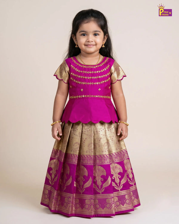 A young girl modeling a traditional Indian outfit consisting of a magenta pink and gold embroidered peplum top with a matching pleated skirt featuring gold zari patterns, set against a plain beige studio background.