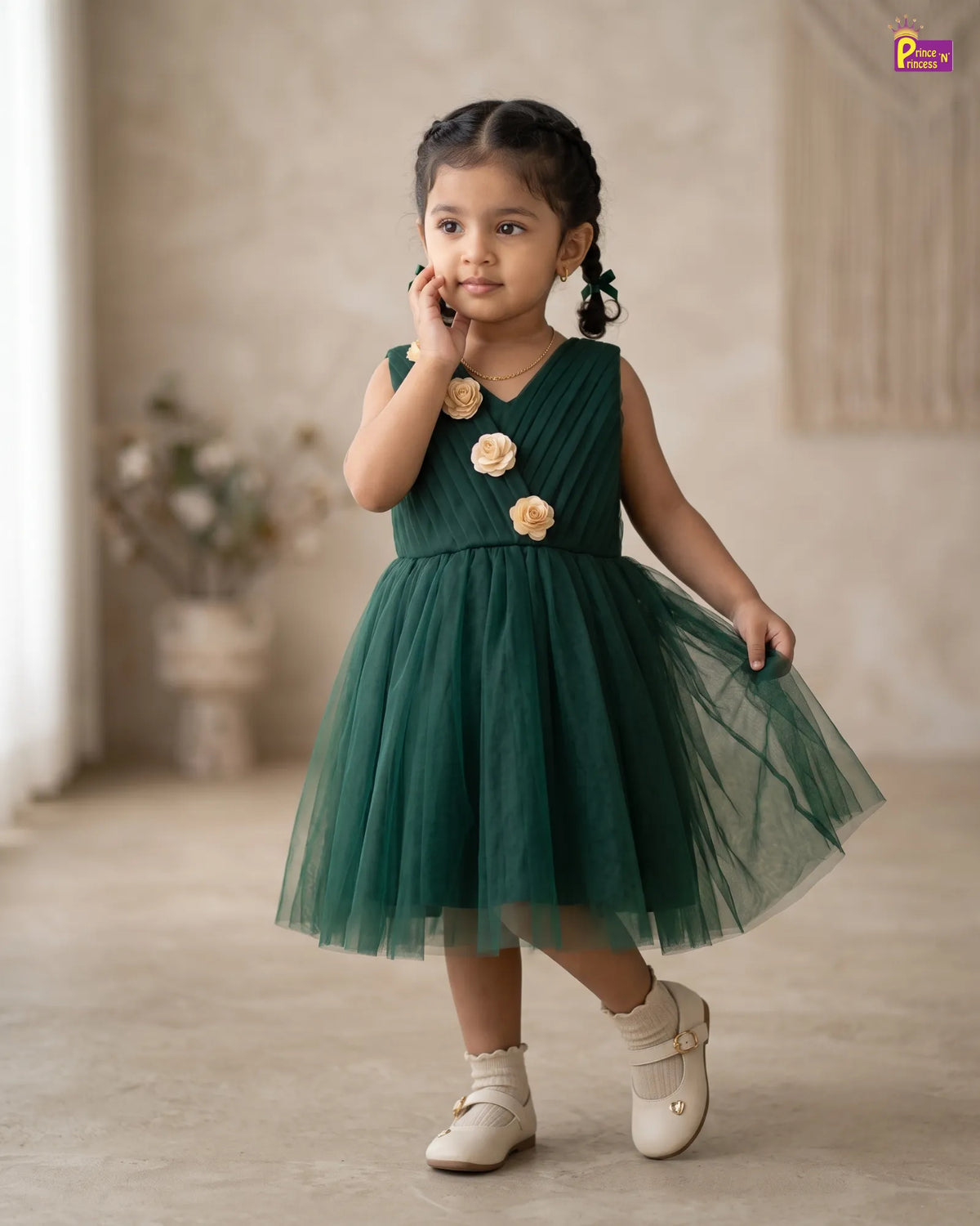 A young girl wearing a sleeveless dark green tulle dress with three beige fabric flowers on the bodice, standing in a softly lit indoor studio setting.