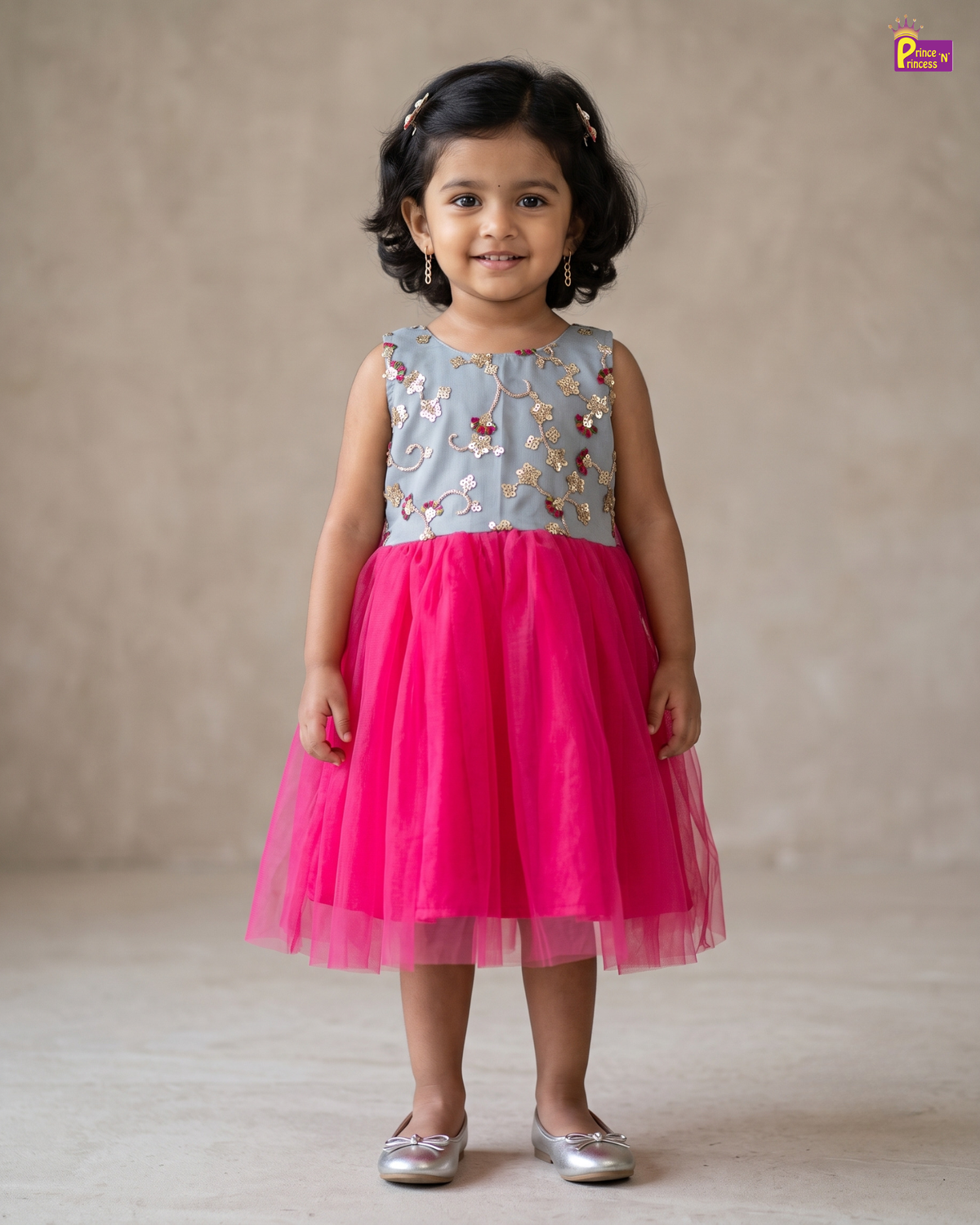 A young girl with dark curly hair is wearing a sleeveless two-piece dress with a light blue-gray top featuring colorful floral embroidery and a bright pink tulle skirt, standing against a neutral beige background.