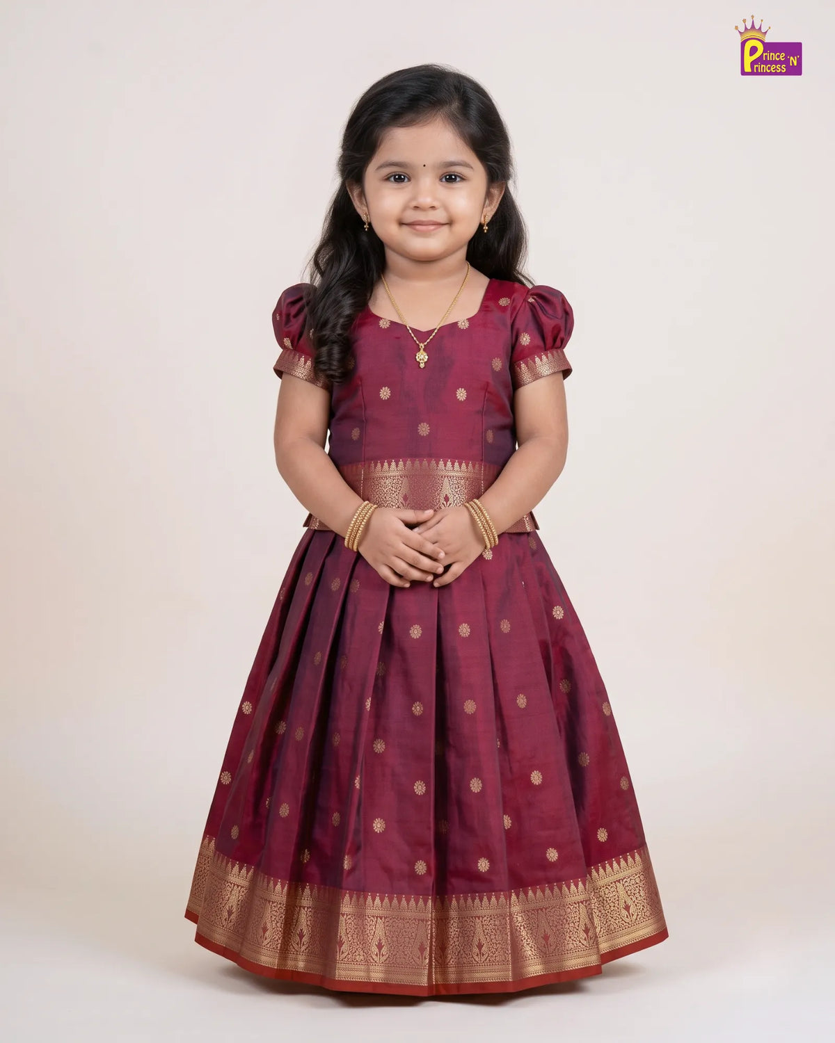A young girl modeling a traditional Indian dress, featuring a deep maroon skirt with gold polka dots and a wide gold zari border, paired with a matching maroon blouse. She is standing against a plain off-white studio background.