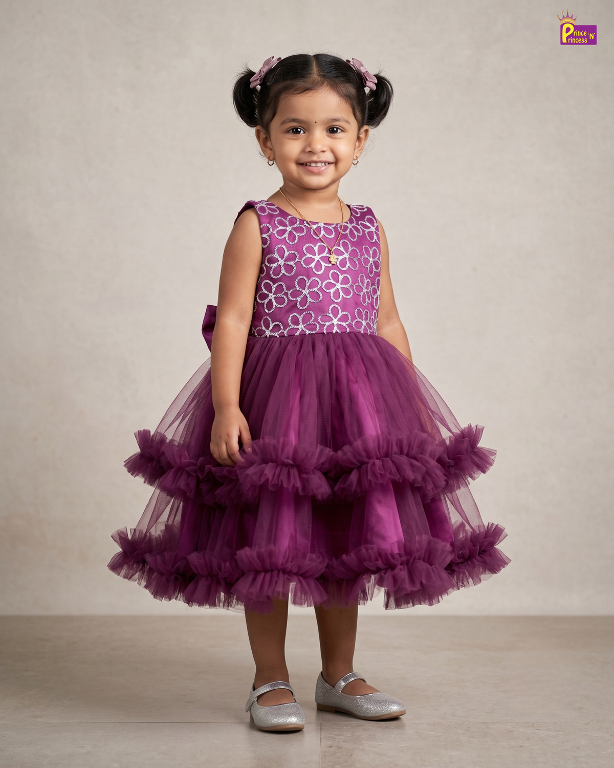 A young girl with her hair in two buns stands wearing a sleeveless purple dress with a white floral pattern on the bodice and a tiered tulle skirt. She is wearing silver ballet flats against a neutral beige studio background.