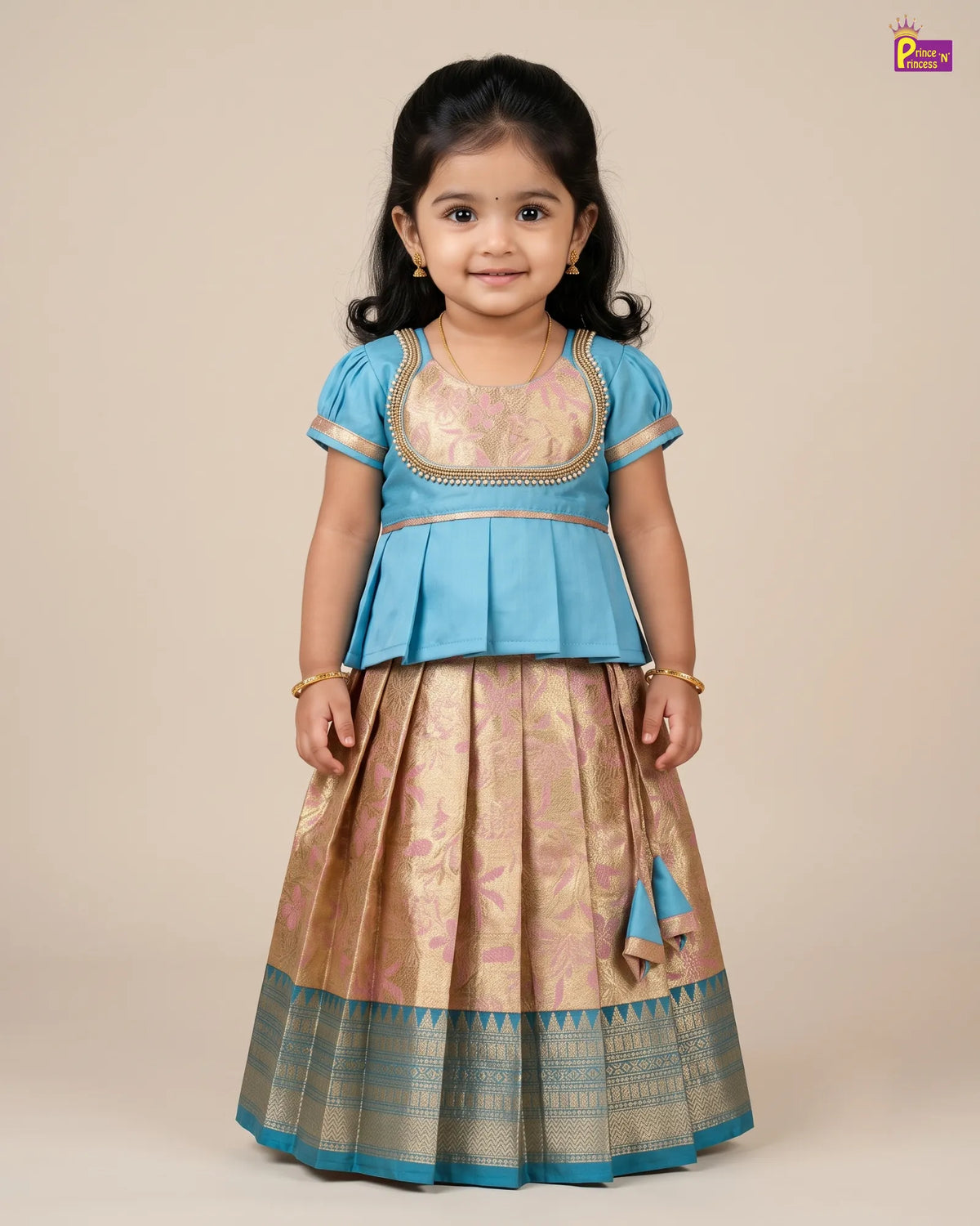 A young girl modeling a traditional Indian ethnic outfit consisting of a light blue peplum blouse with gold embroidery and a matching beige and gold patterned skirt with a blue border, set against a plain beige studio background.