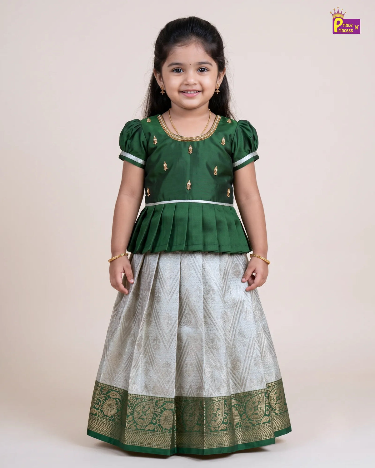 A young girl modeling a traditional Indian outfit consisting of a dark green peplum top with gold embroidery and a light silver-gray patterned skirt with a green border, set against a plain beige background.