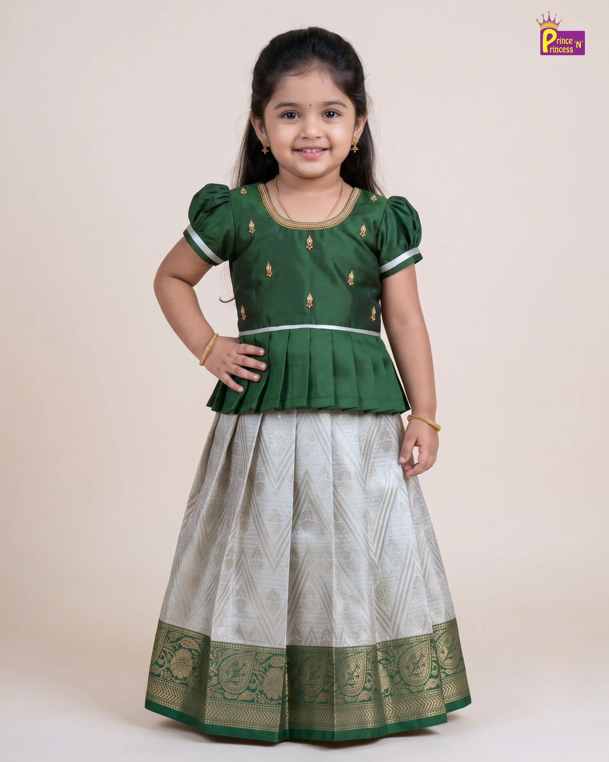 A young girl modeling a traditional Indian ethnic outfit consisting of a dark green peplum top with gold embroidery and a silver patterned skirt with a green border, set against a plain beige background.