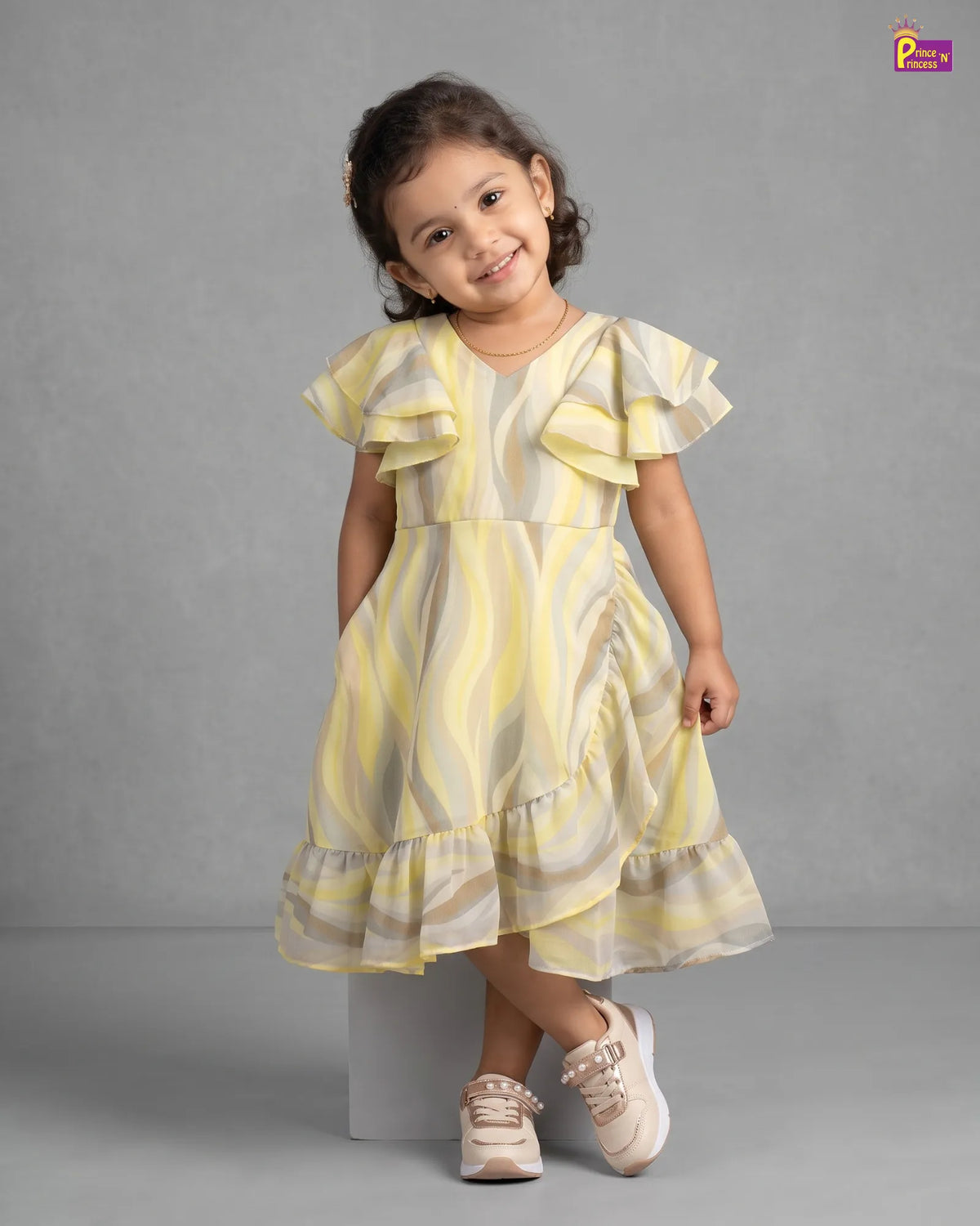 A young girl with dark curly hair stands on a gray studio background wearing a yellow and beige wavy-patterned ruffled dress with beige sneakers.