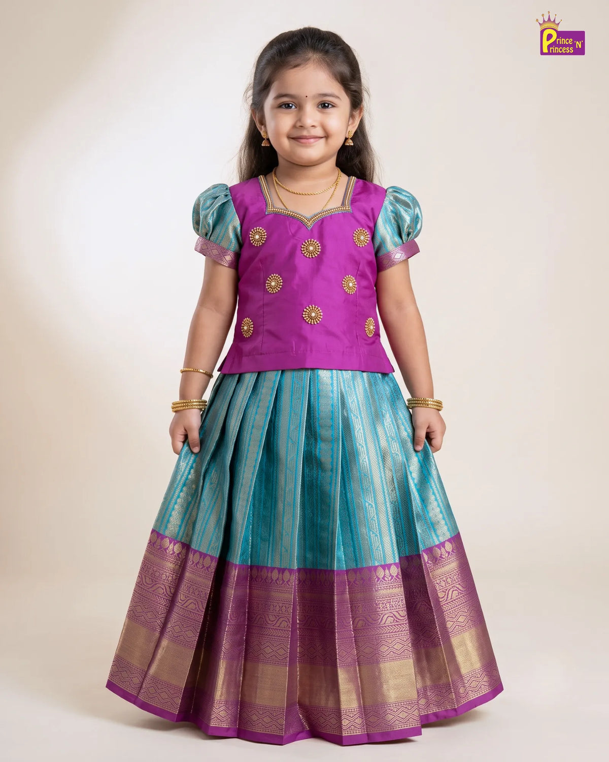A young girl modeling a traditional Indian ethnic dress, featuring a bright purple bodice with gold embroidery and a teal blue skirt with gold zari stripes and a purple border, set against a plain beige studio background.