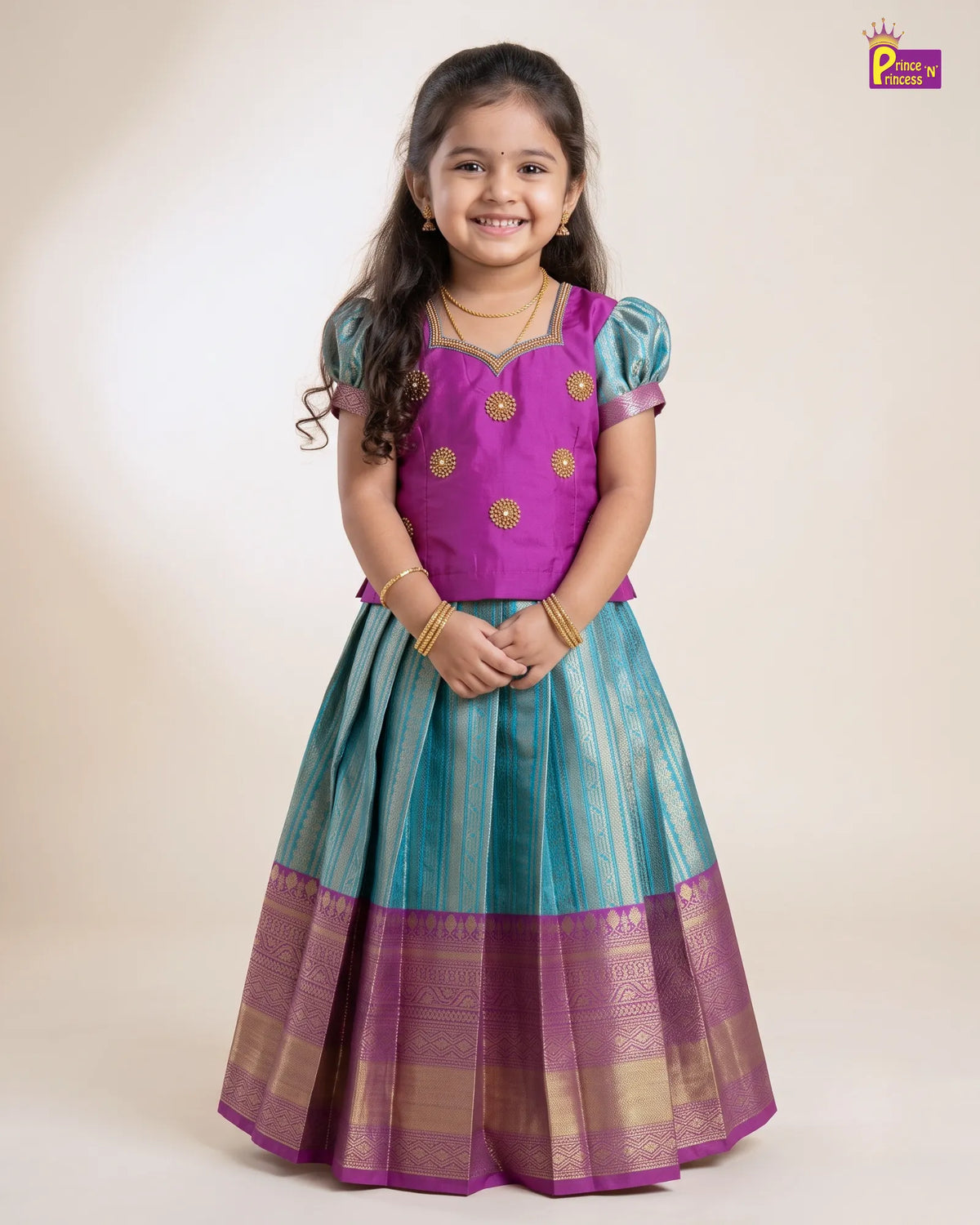 A young girl modeling a traditional Indian lehenga choli dress. The dress features a vibrant purple choli with gold embroidered details, and a pleated skirt in teal blue and purple with gold zari stripes and borders.