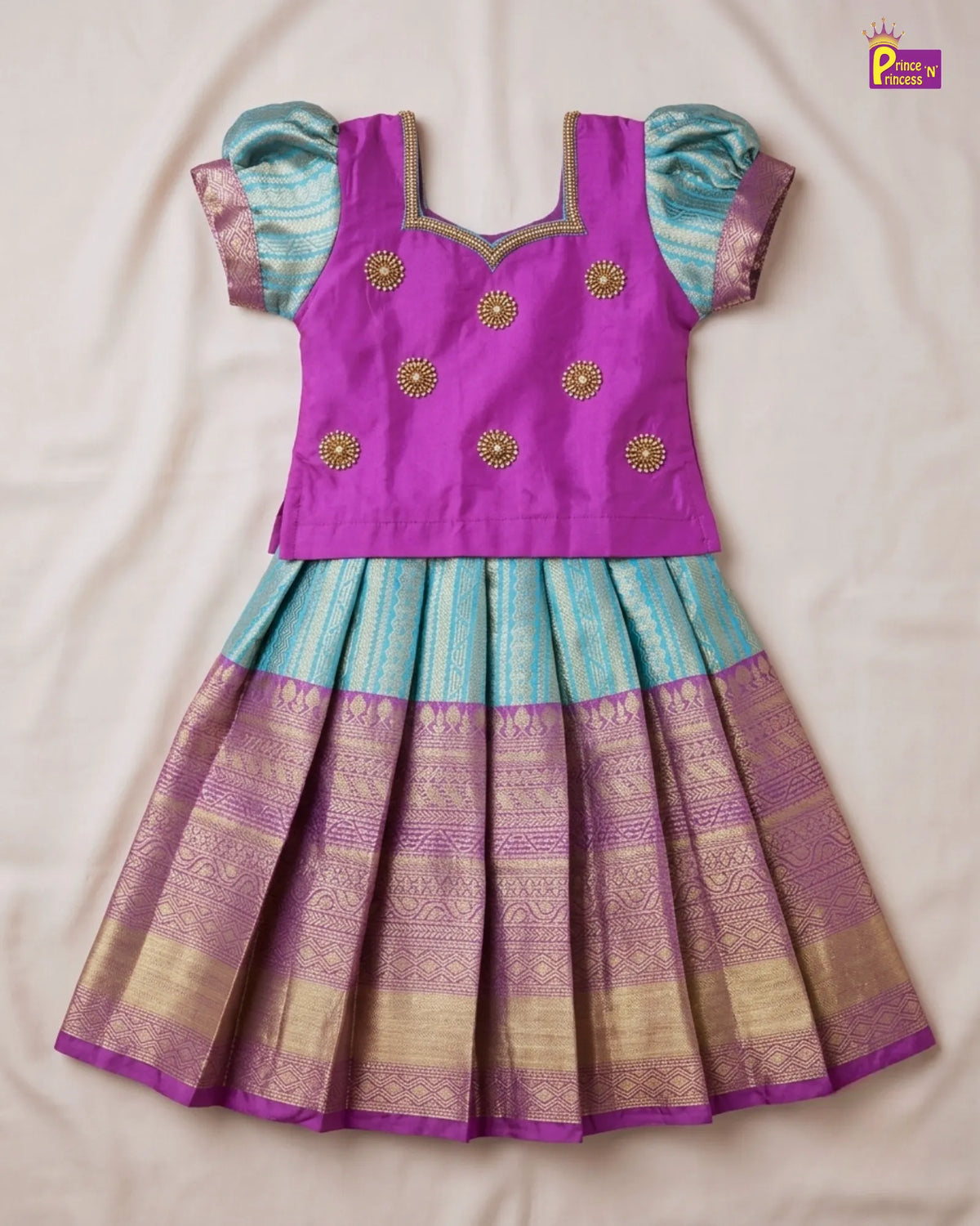 A traditional Indian girl's dress laid flat on a light beige fabric background. The dress features a bright purple sleeveless bodice with gold embroidered motifs, light blue puff sleeves, and a pleated skirt with a purple and gold patterned Banarasi fabric.