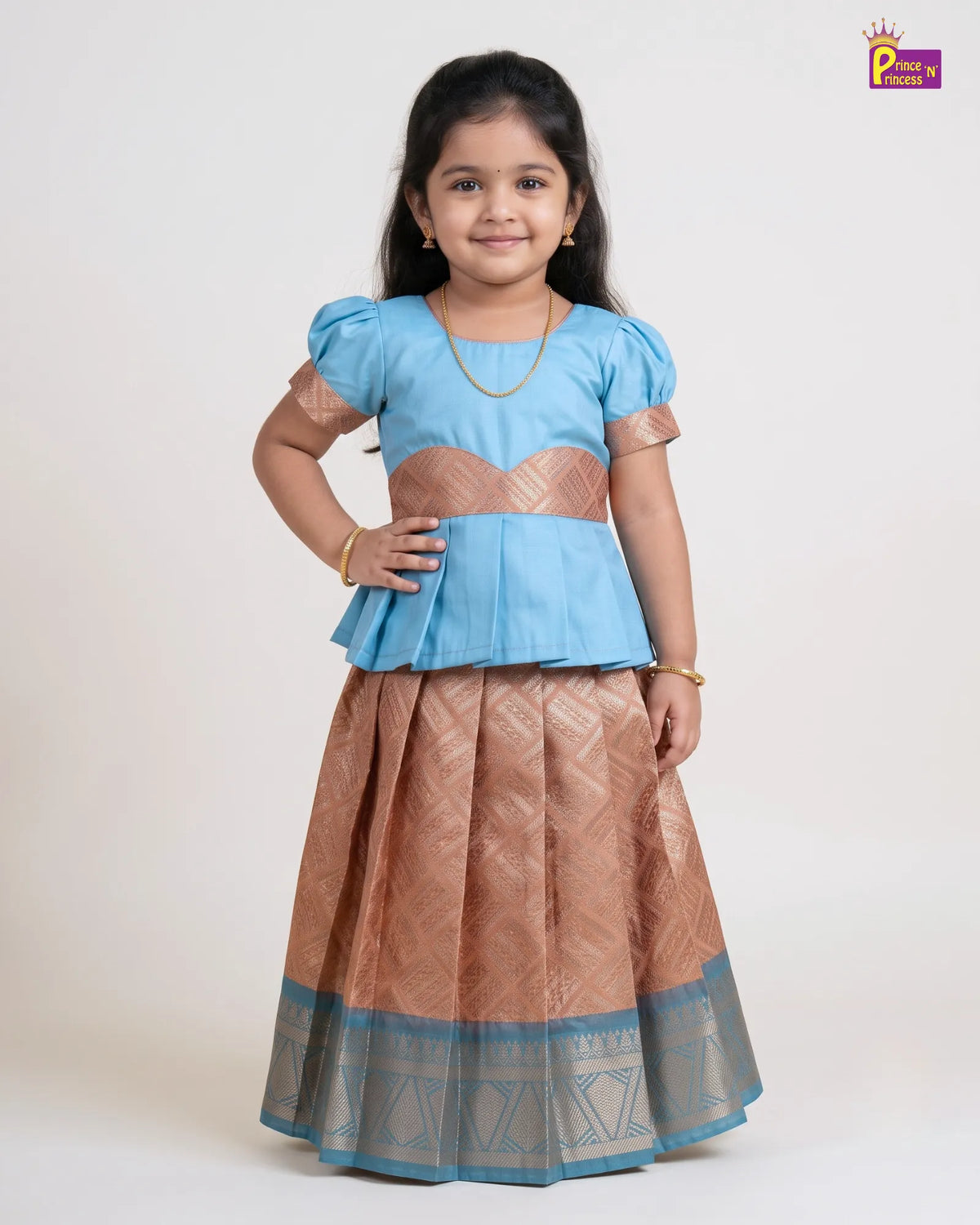 A young girl modeling a traditional Indian outfit consisting of a light blue peplum top and a bronze-brown flared skirt with a blue border, shot against a plain light background.