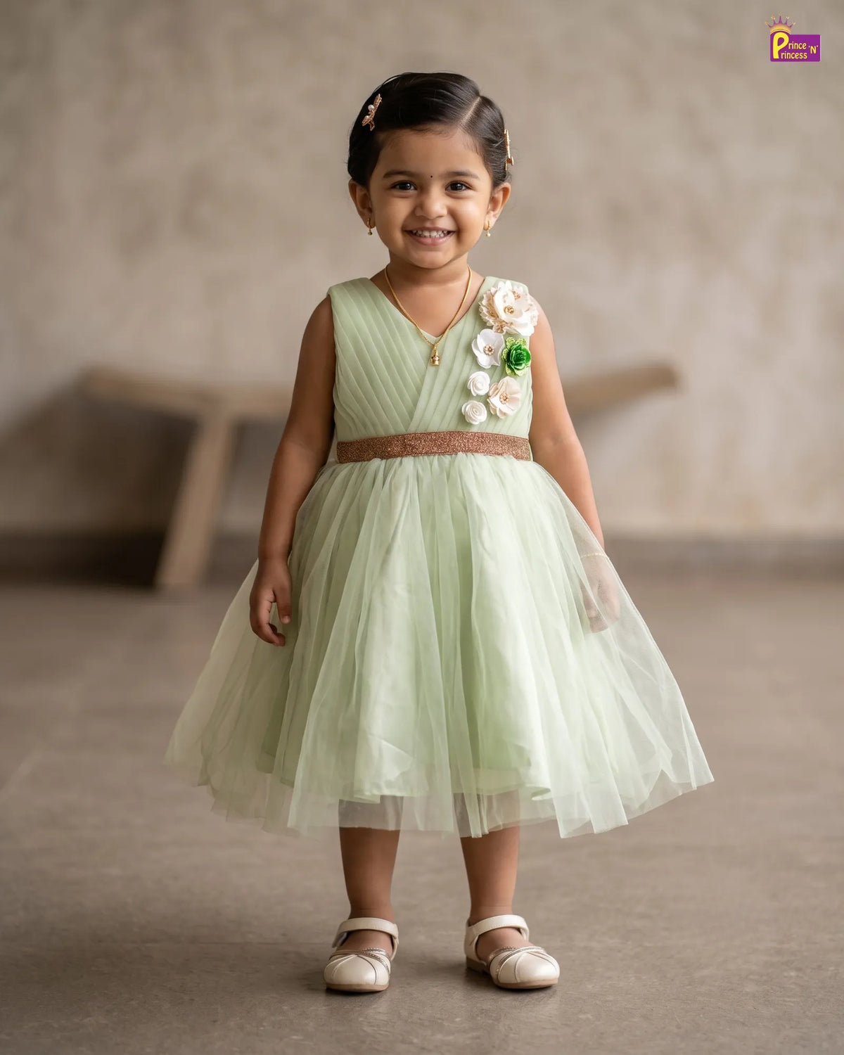 A young girl stands smiling in a sleeveless mint green tulle dress with a brown sash and white floral embellishments on the chest, photographed indoors against a softly blurred neutral background.