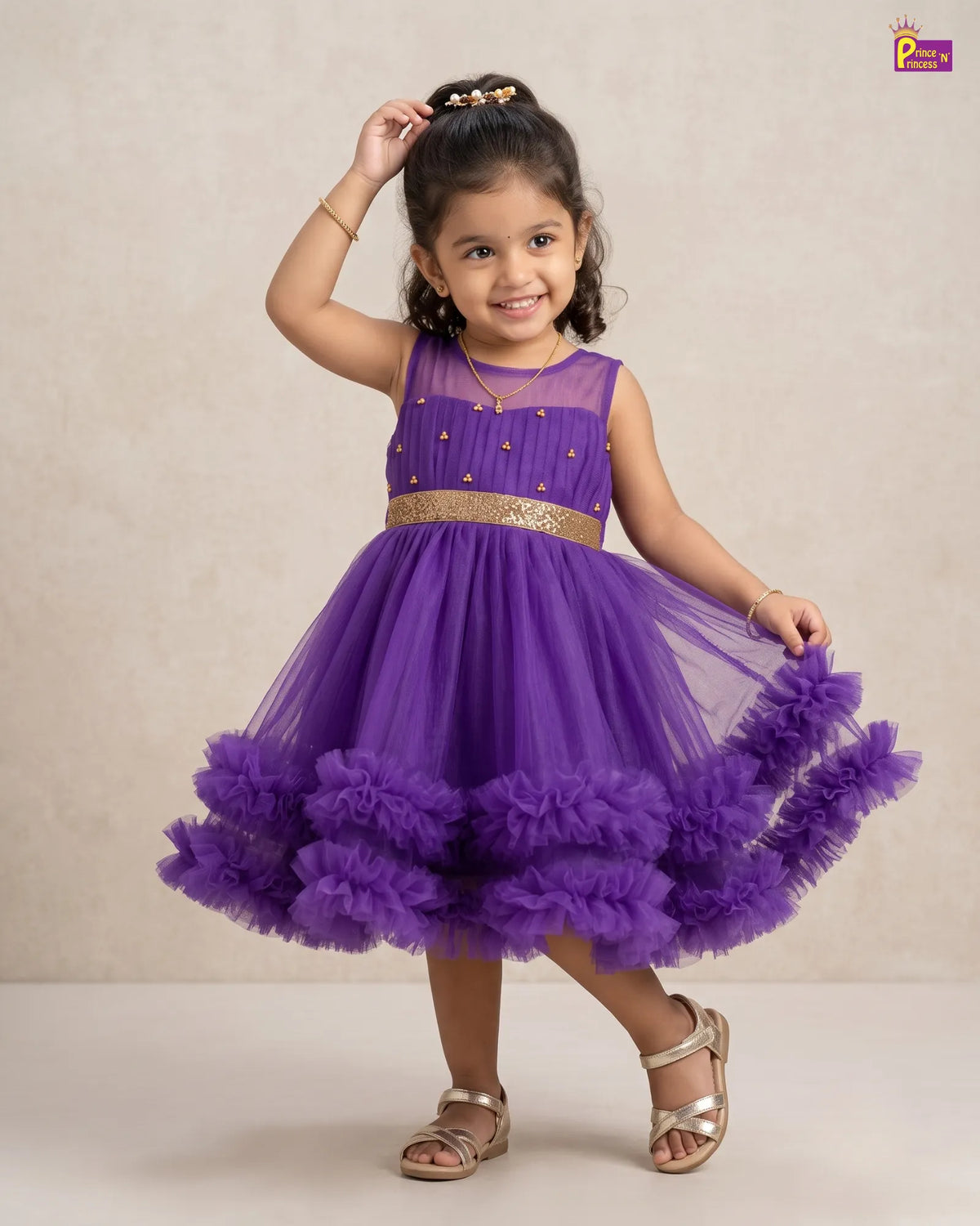 A young girl modeling a sleeveless purple tulle dress with a ruffled skirt, gold embellishments, and a gold waistband, standing against a neutral beige background.