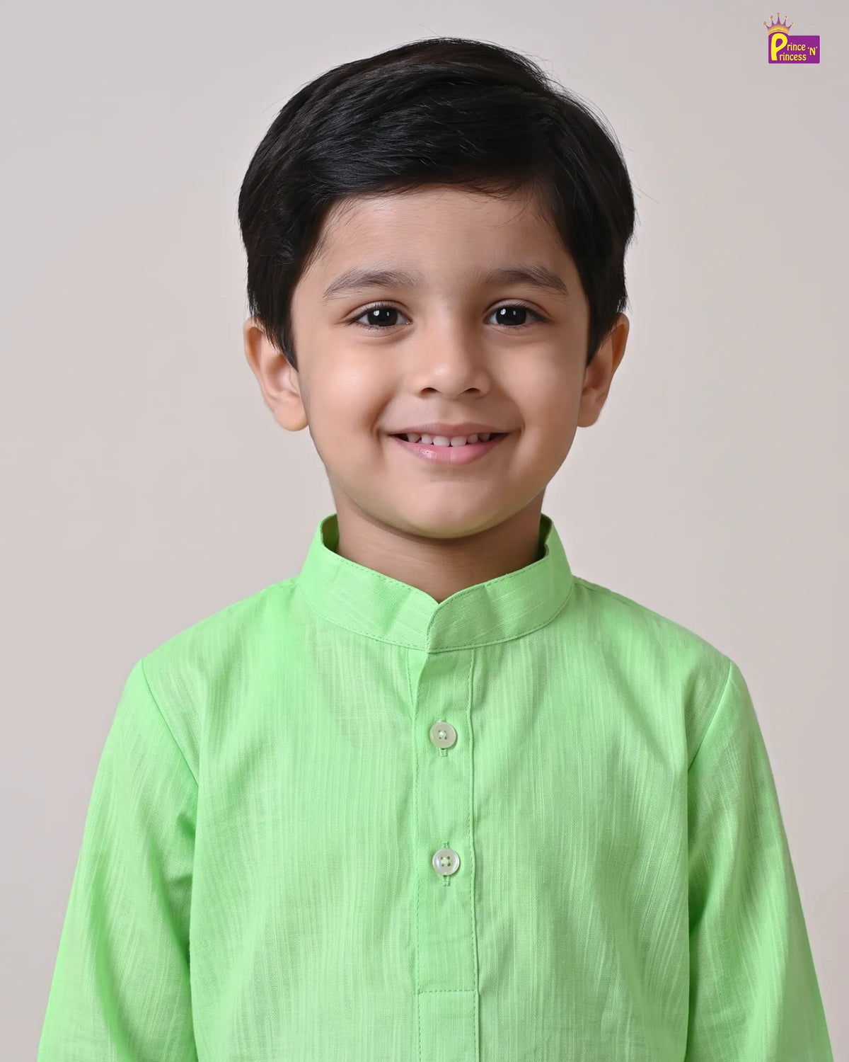 A young boy with dark hair smiling, wearing a light green long-sleeve kurta with a mandarin collar and silver buttons, set against a plain light beige background.