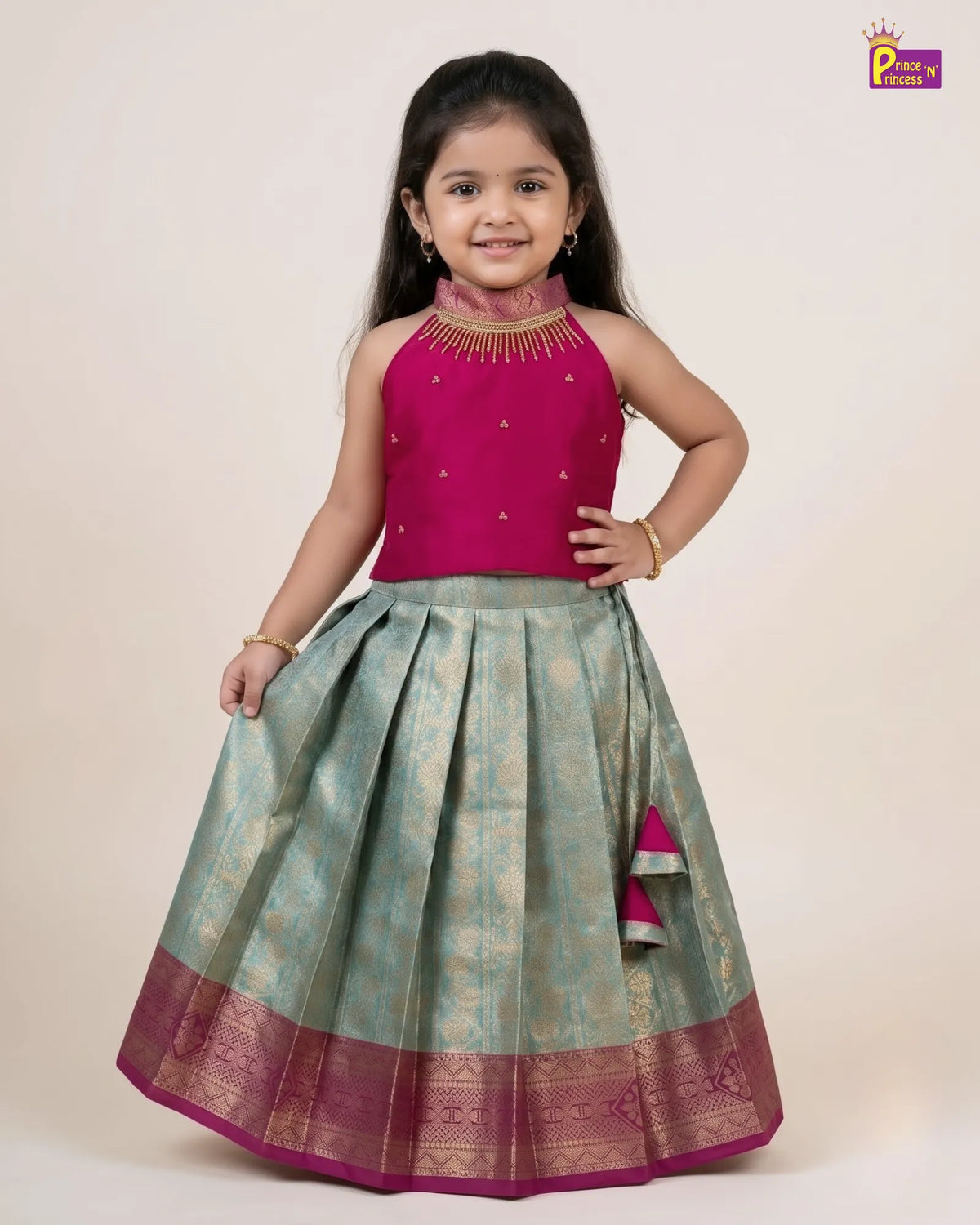 A young girl modeling a traditional Indian ethnic wear set consisting of a magenta pink sleeveless top with gold embroidery and a matching pleated light green skirt with a magenta border, on a plain beige background.