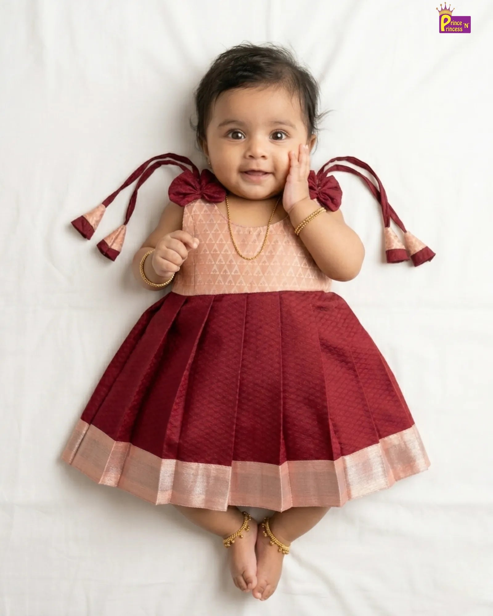 Baby wearing a maroon and peach dress with tassels on a white background