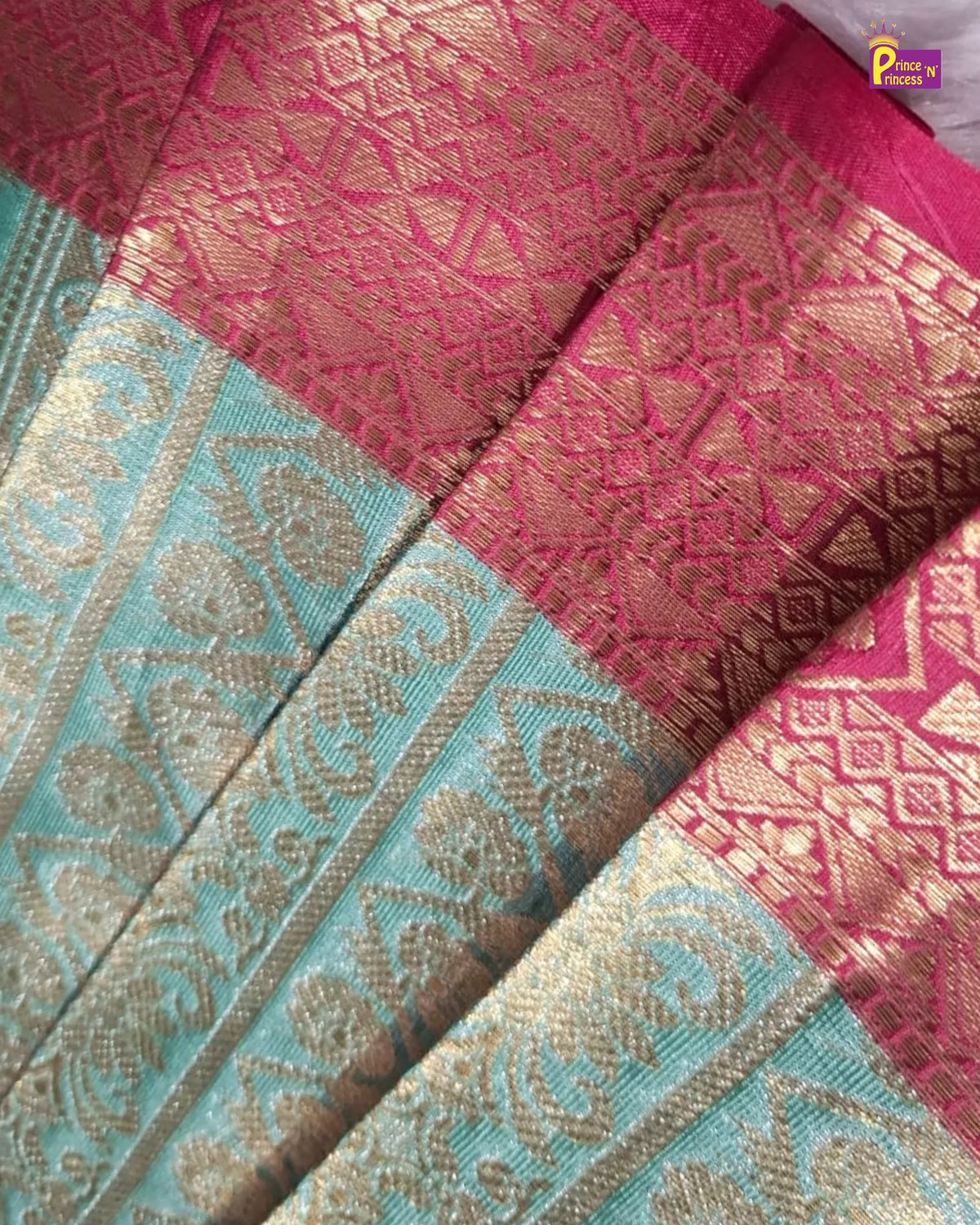 A close-up of a woven textile, likely a sari, featuring a vibrant pink and light blue body with intricate gold zari metallic patterns and borders.