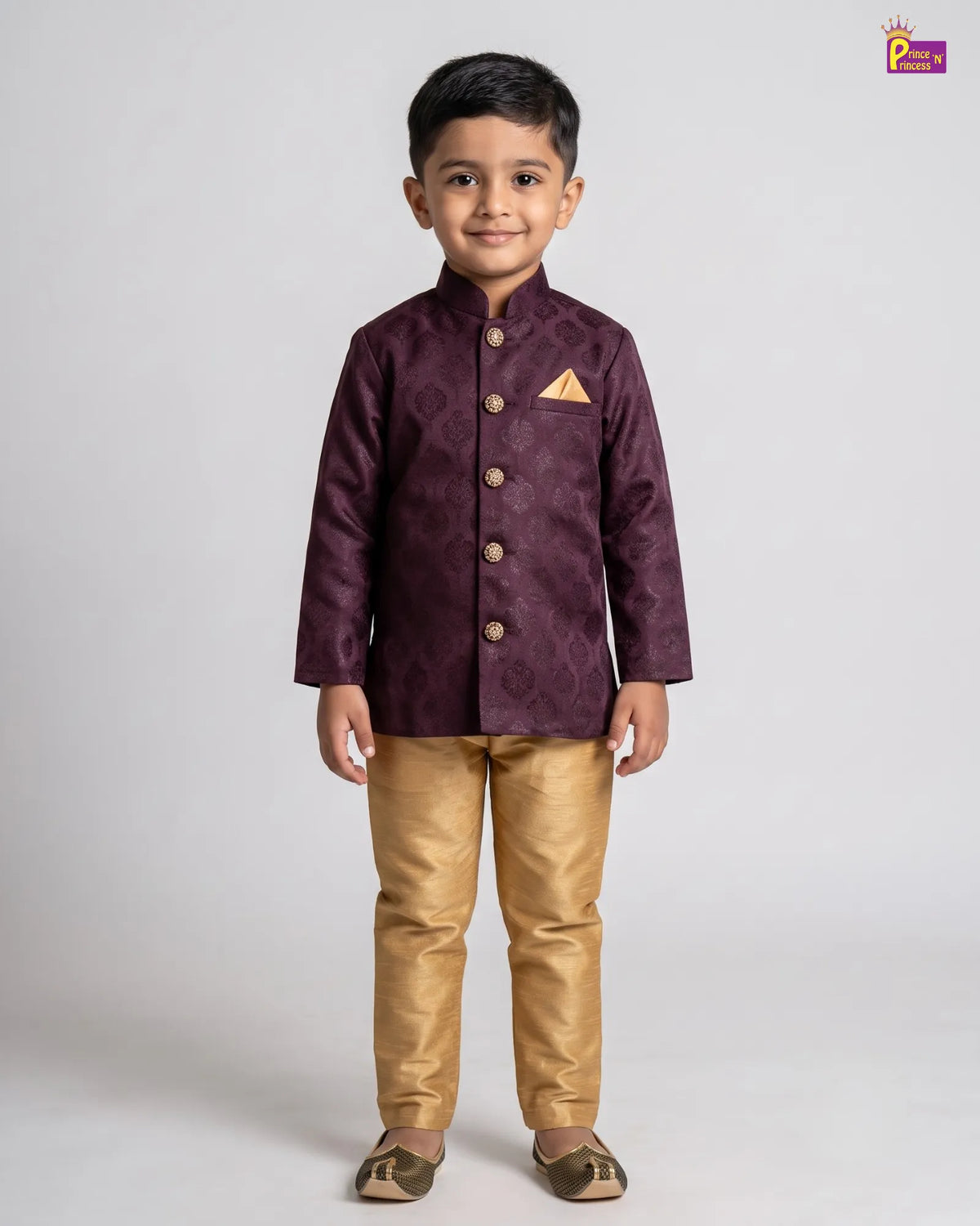 A young boy modeling a traditional Indian outfit consisting of a deep purple embroidered sherwani jacket with gold buttons and a matching gold pocket square, paired with metallic gold trousers and brown shoes, against a light gray studio background.