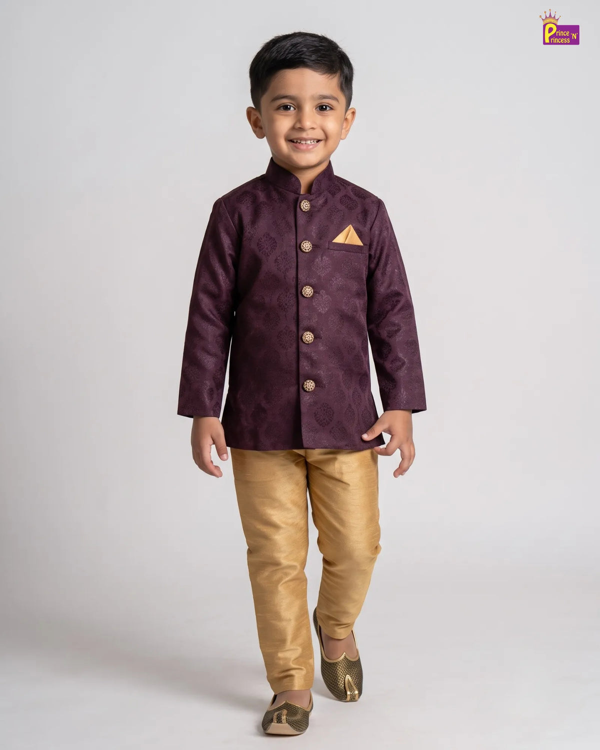 A young boy modeling a traditional Indian outfit consisting of a deep purple textured sherwani jacket with gold buttons and a matching pocket square, paired with shiny gold trousers and gold shoes, against a light gray studio background.