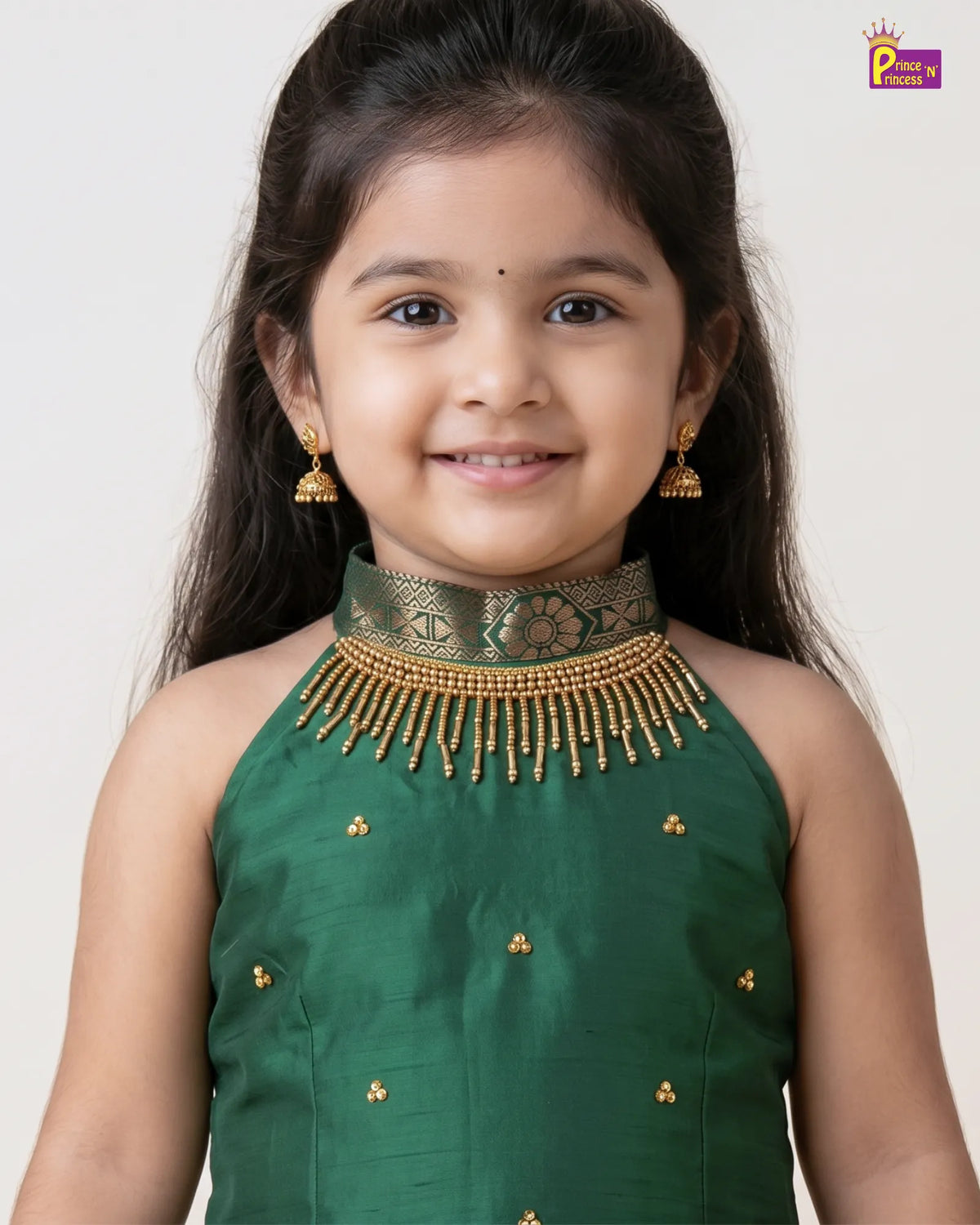 A young girl with dark hair is wearing a sleeveless dark green traditional top with a decorative gold beaded collar and matching gold earrings, photographed against a plain light background.