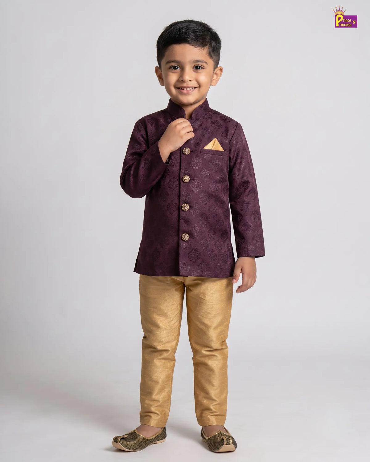 A young boy modeling a traditional Indian outfit consisting of a deep purple embroidered jacket with gold buttons and a matching pocket square, paired with metallic gold trousers and brown shoes, against a plain light gray background.