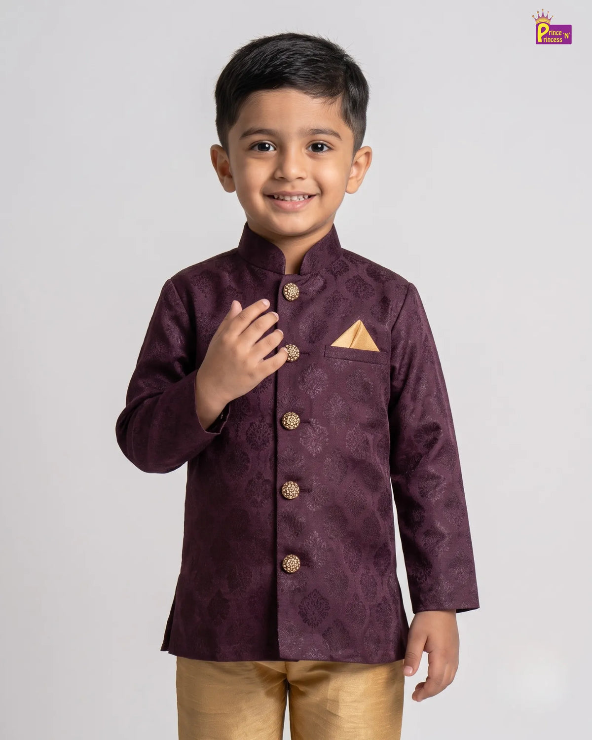 A young boy modeling a dark purple textured sherwani jacket with gold buttons and a gold pocket square, paired with gold trousers, against a light gray background.