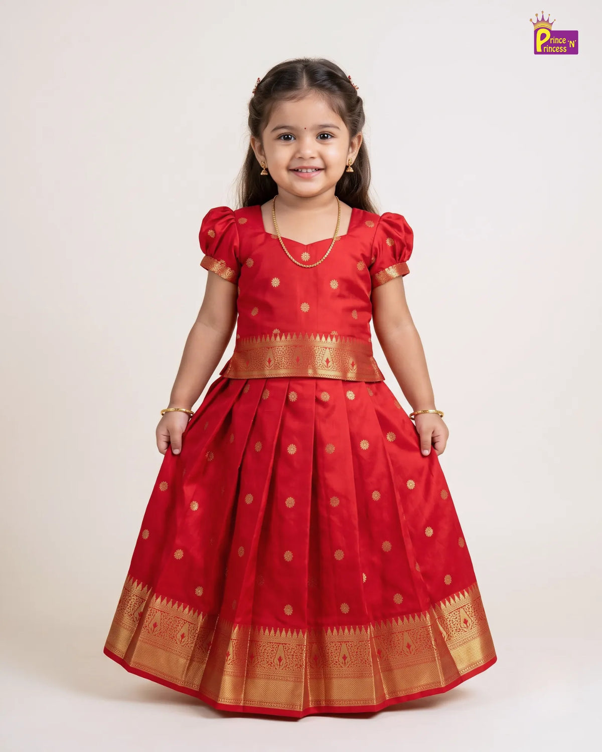 A young girl modeling a traditional red Indian ethnic dress with gold motifs and a flared skirt, photographed against a plain light beige studio background.