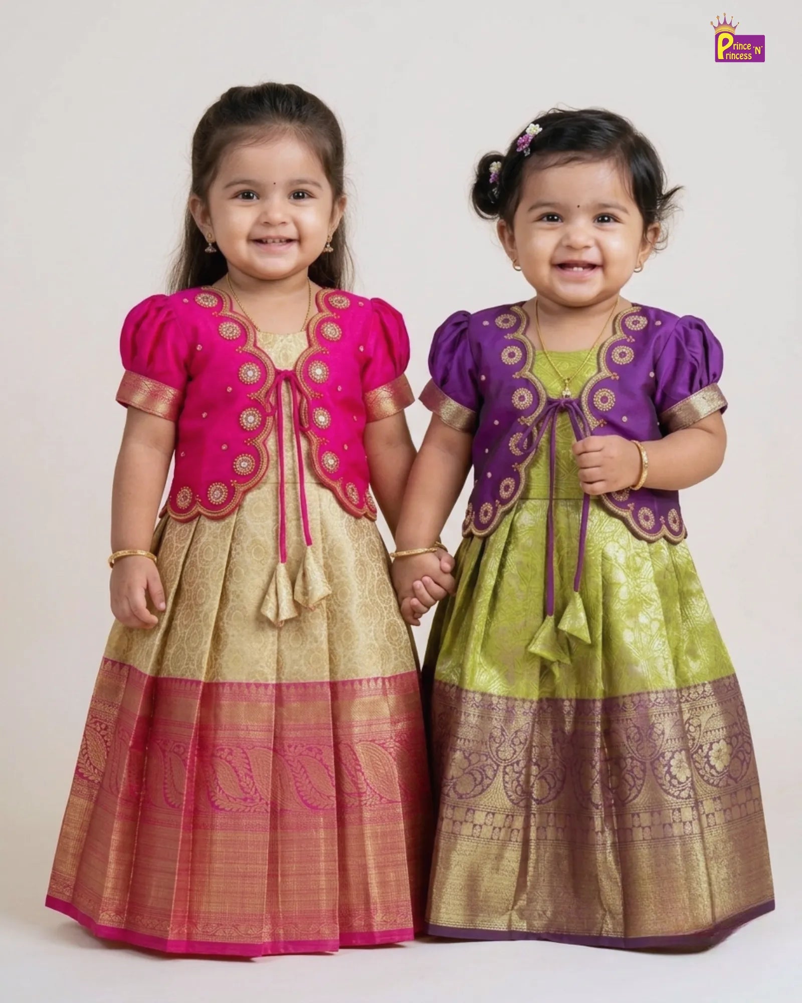 Two young girls modeling traditional Indian ethnic wear, standing side-by-side and holding hands. The girl on the left wears a pink kurta with gold embroidery over a beige and pink Banarasi skirt.