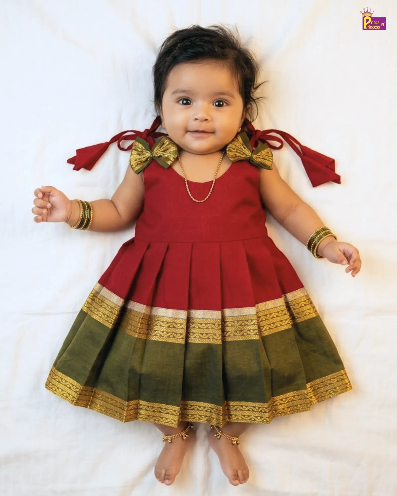 Baby wearing a red and green traditional outfit with gold accents on a white background