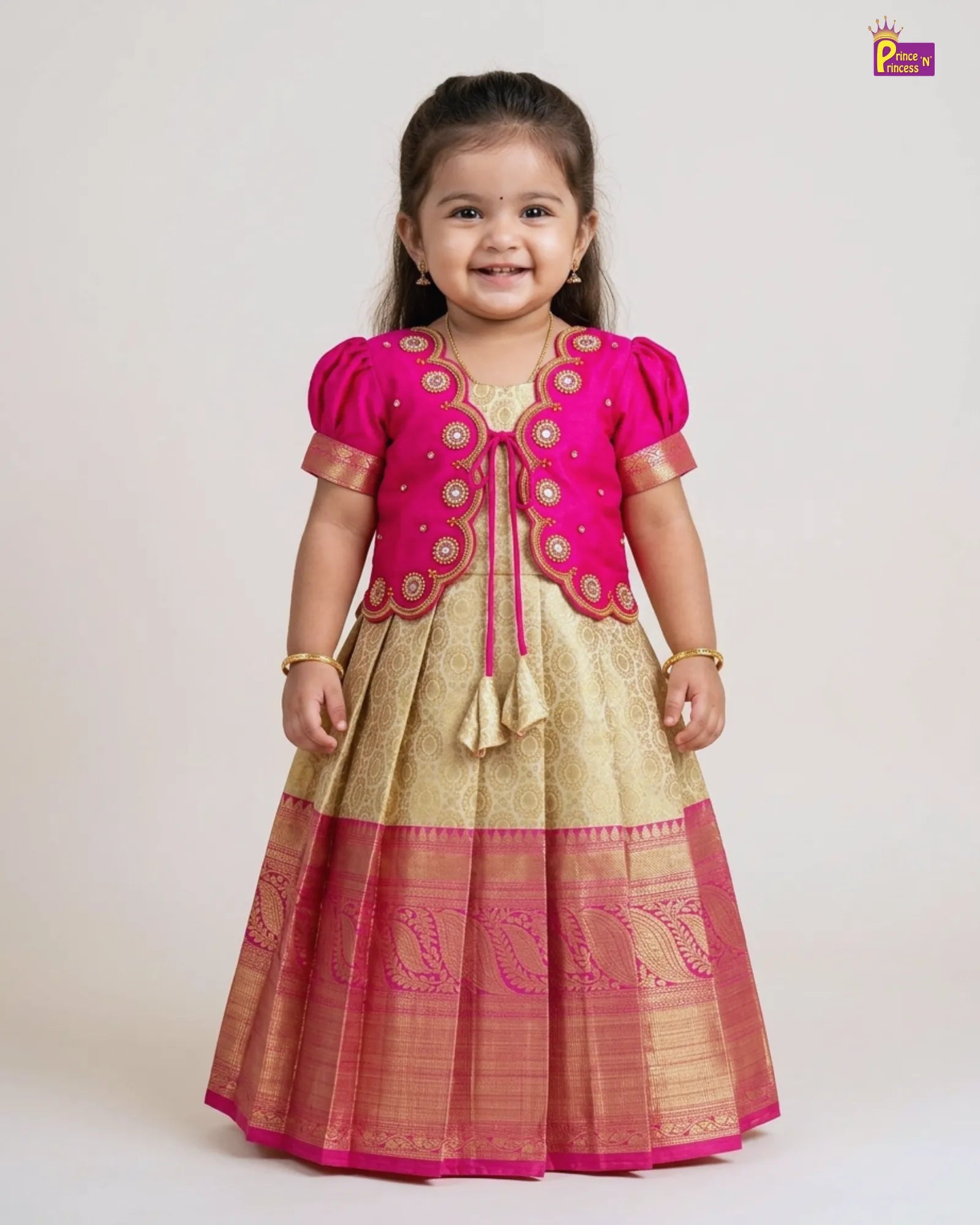 A young girl modeling a traditional Indian ethnic outfit consisting of a bright pink embroidered jacket and a beige skirt with a pink border, photographed against a plain light gray studio background.