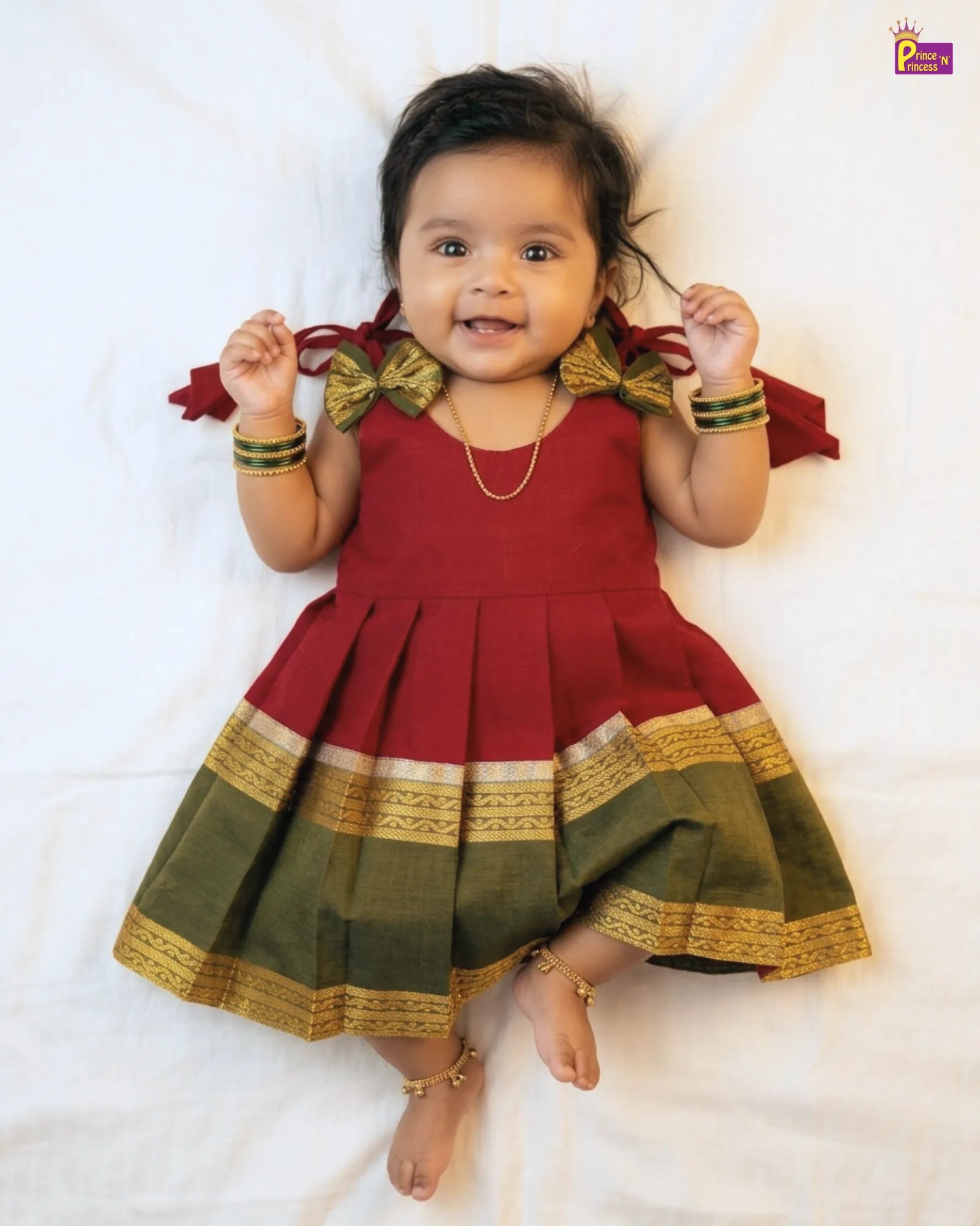 Baby wearing a red and green traditional outfit with gold accents on a white background