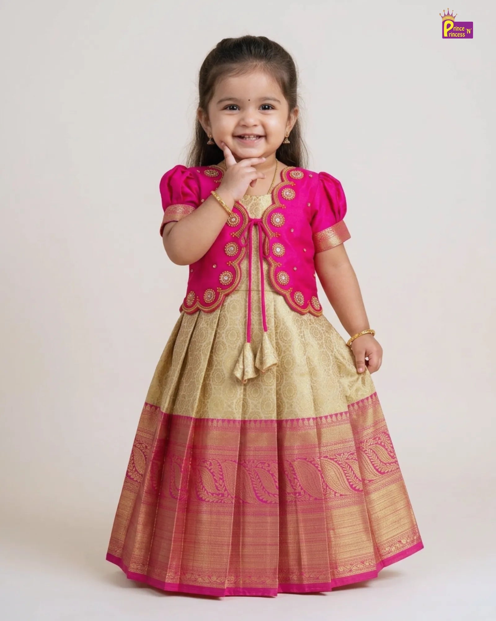 A young girl modeling a traditional Indian dress with a bright pink embroidered bodice and a beige flared skirt with a pink border, photographed against a plain light background.