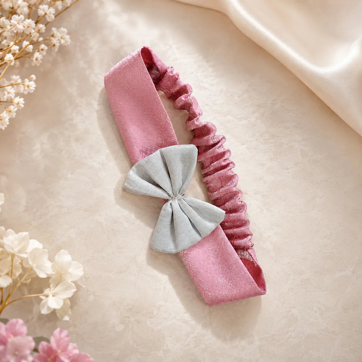 A pink satin and elastic headband with a light gray bow, styled on a beige textured surface with dried flowers and a cream satin fabric in the background.