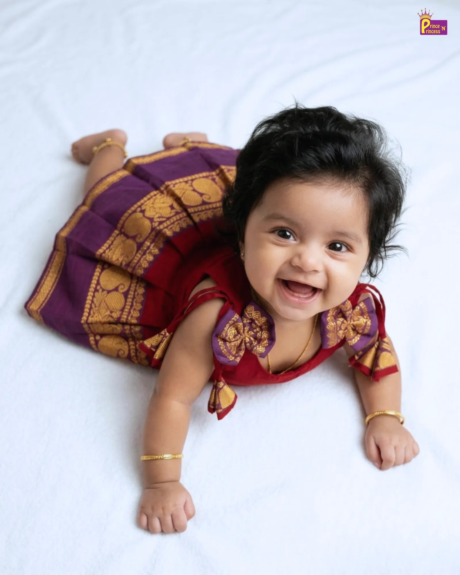 Baby wearing a traditional outfit with a purple and gold top and red dress, lying on a white background.