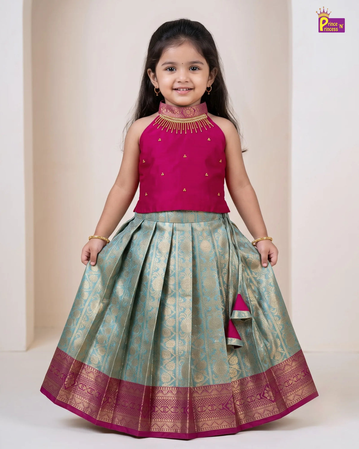 A young girl modeling a traditional Indian ethnic dress consisting of a magenta sleeveless top with gold embroidery and a matching pleated light blue-green skirt with gold zari patterns and a magenta border.