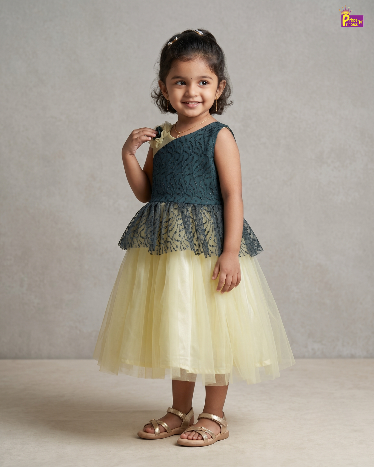 A young girl modeling a sleeveless two-tone dress with a dark teal lace bodice and a pale yellow tulle skirt, paired with gold sandals, set against a neutral textured studio backdrop.