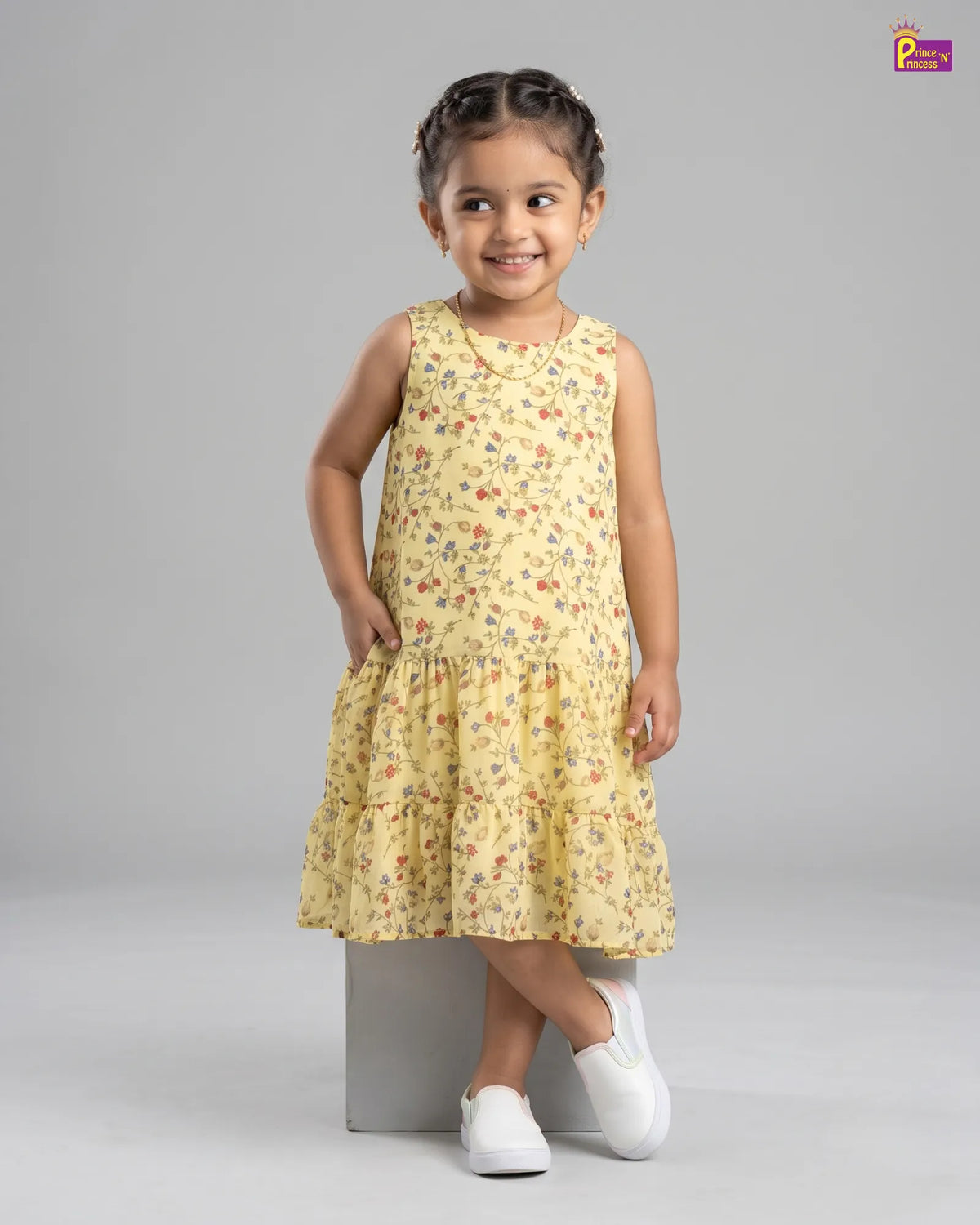 A young girl with braided hair stands on a gray block wearing a sleeveless yellow tiered dress with a small multicolor floral print and white slip-on shoes, photographed against a light gray studio background.