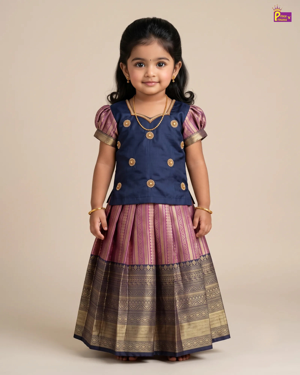 A young girl modeling a traditional Indian outfit consisting of a navy blue top with gold buttons and a pleated skirt in purple and gold zari fabric, set against a plain beige background.