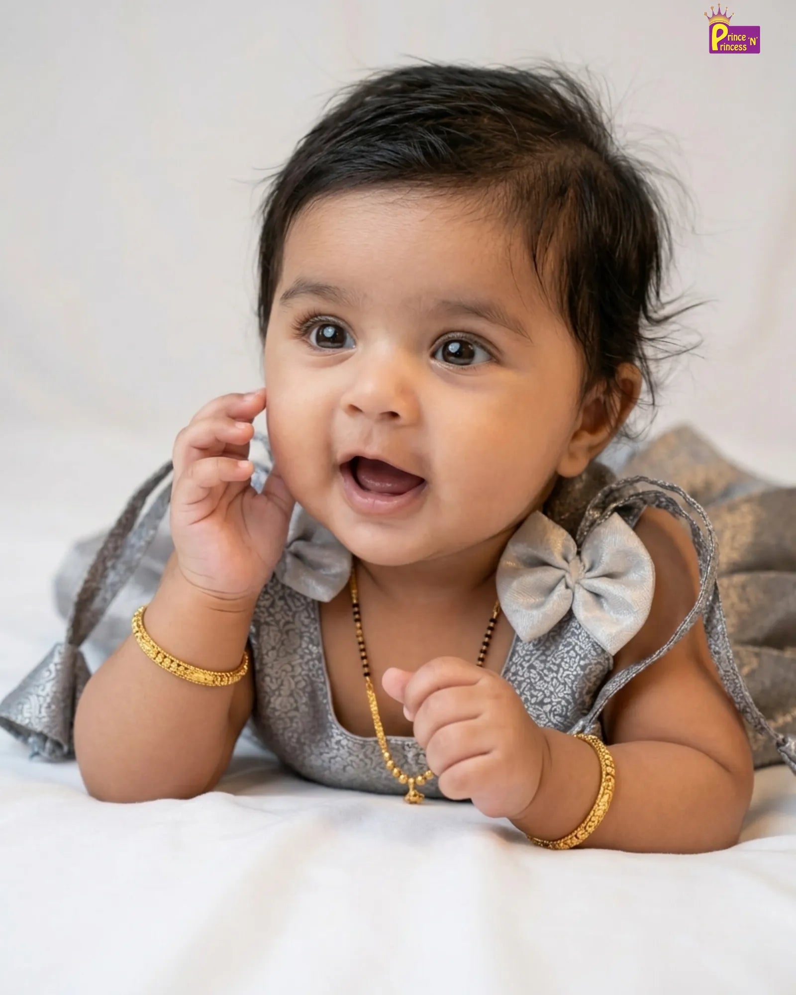 Baby wearing a silver dress with bow details and gold jewelry on a white background