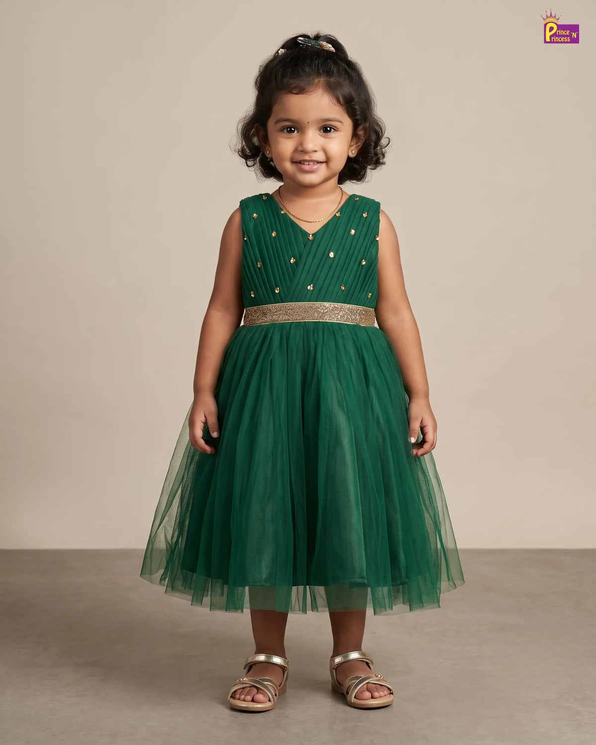 A young girl with dark curly hair wearing a sleeveless emerald green party dress with gold polka dots and a metallic gold waistband, standing against a neutral beige studio background.