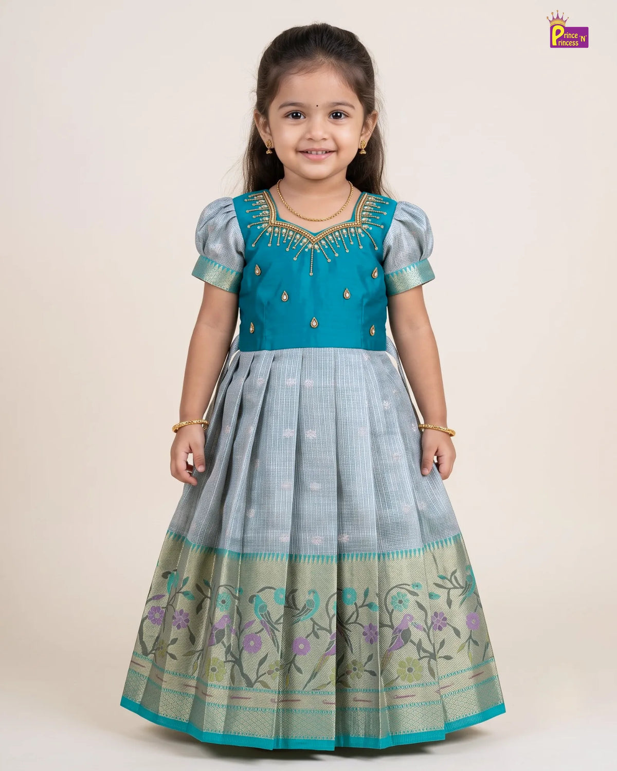 A young girl modeling a traditional Indian dress with a turquoise blue bodice and a light blue-gray flared skirt featuring floral embroidery and a gold border, set against a plain beige studio background.