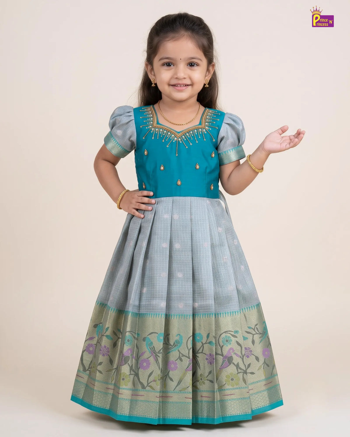 A young girl modeling a traditional Indian salwar kameez dress. The dress features a turquoise blue bodice with gold embroidery, short light blue sleeves, and a light gray-blue flared skirt with a floral border.