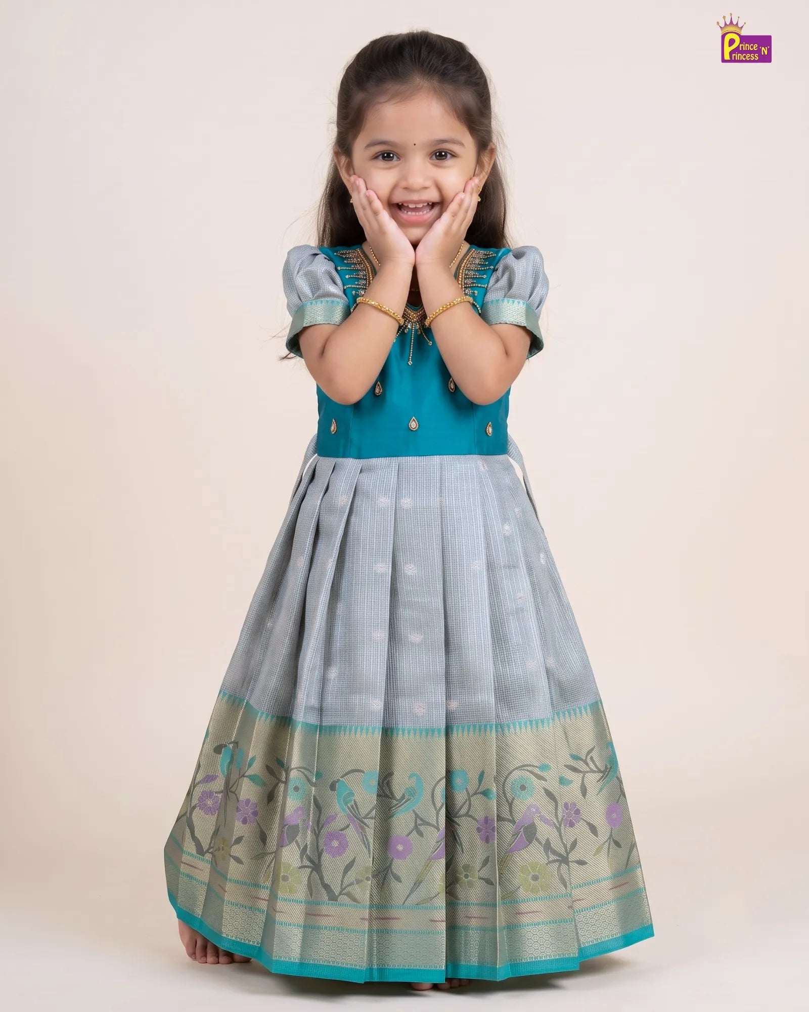 A young girl modeling a traditional Indian dress, featuring a teal blue bodice with gold embroidery and a pleated light blue skirt with a floral border, set against a plain beige background.