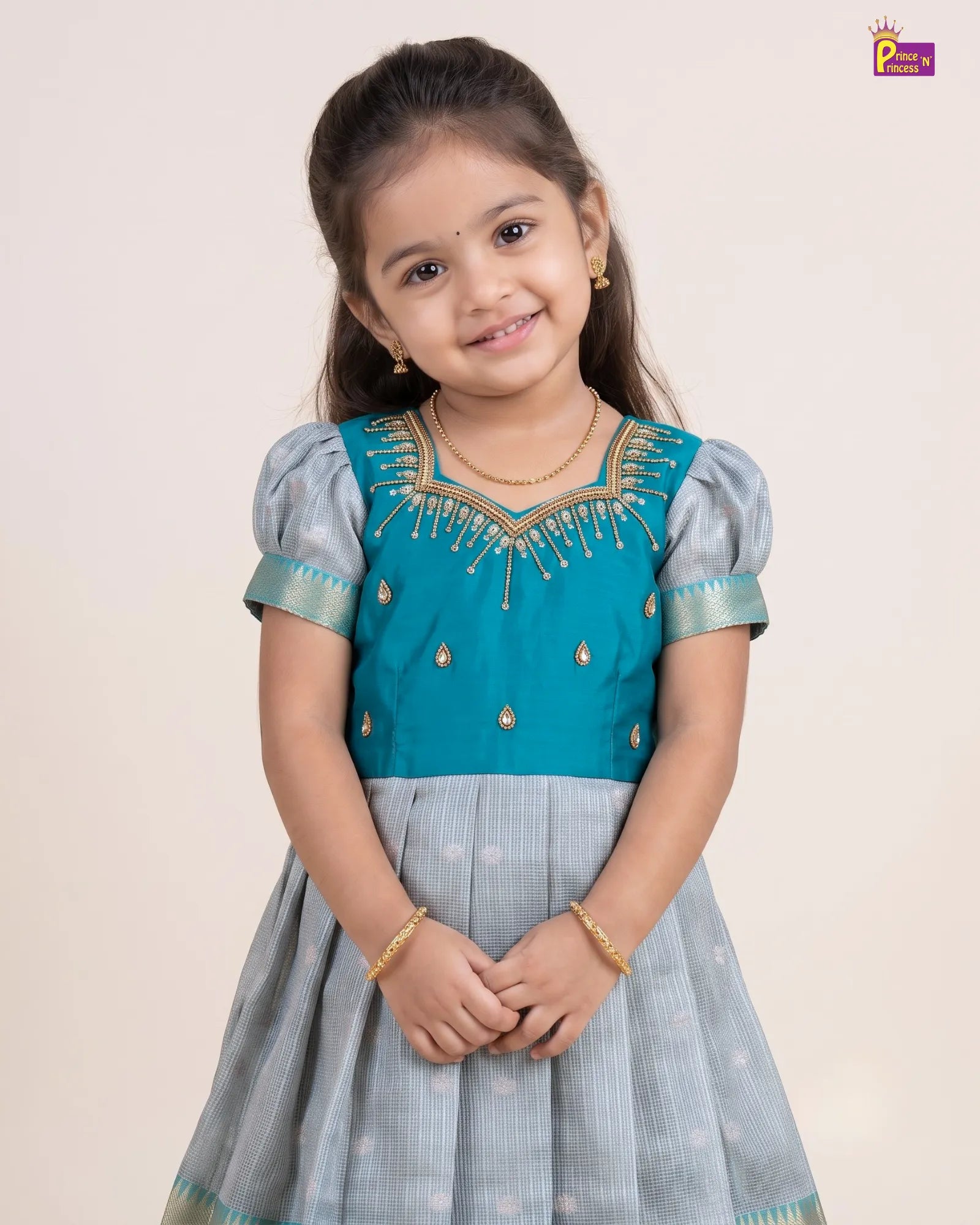 A young girl modeling a traditional Indian dress with a teal blue bodice and a light gray-blue skirt, featuring gold embroidery and puffed sleeves, set against a plain beige background.