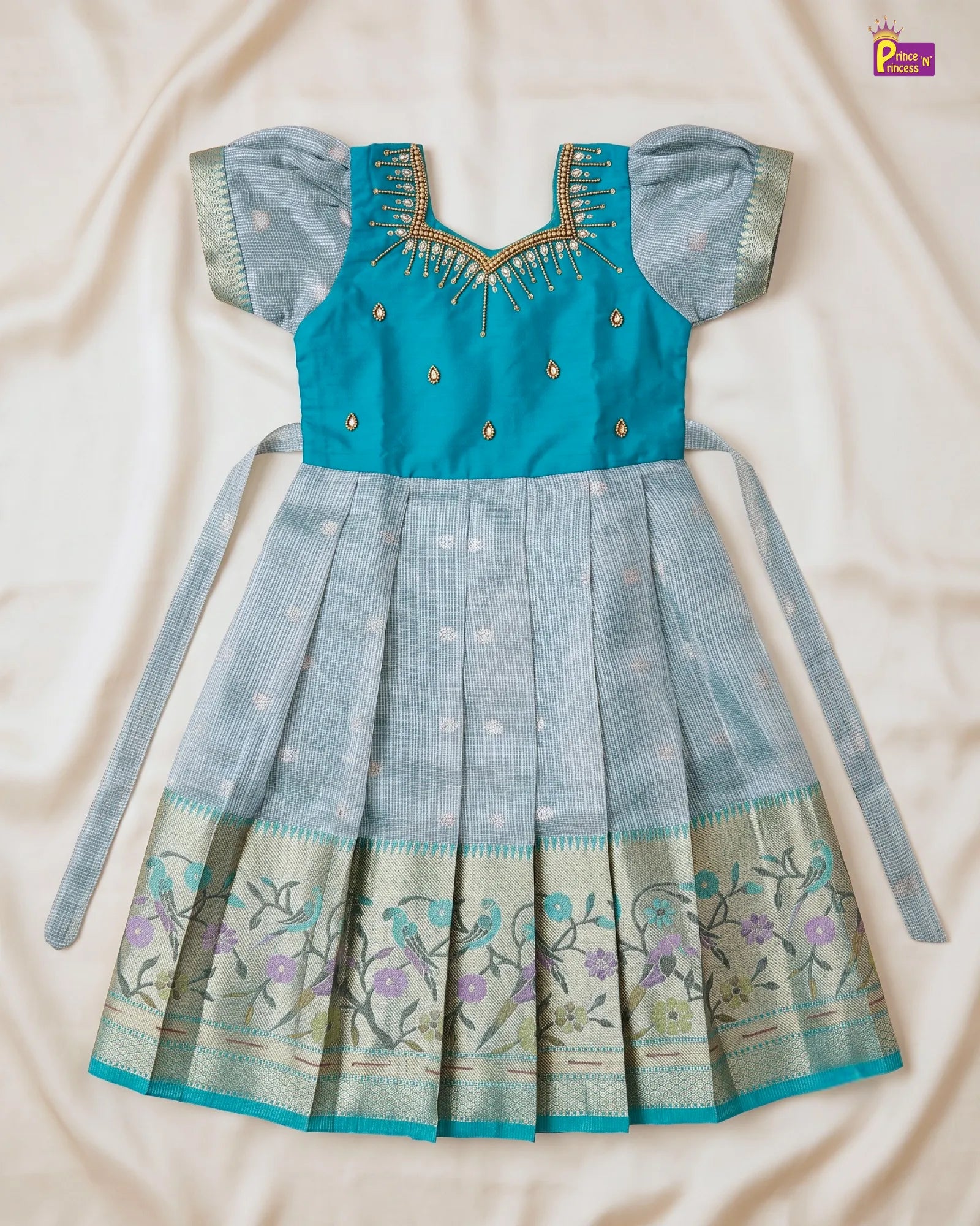 A traditional Indian girl's dress (Anarkali) laid flat on a beige fabric background. The dress features a vibrant turquoise blue bodice with gold embroidery, contrasting with a light blue pleated skirt decorated with a floral border and tie-strings.