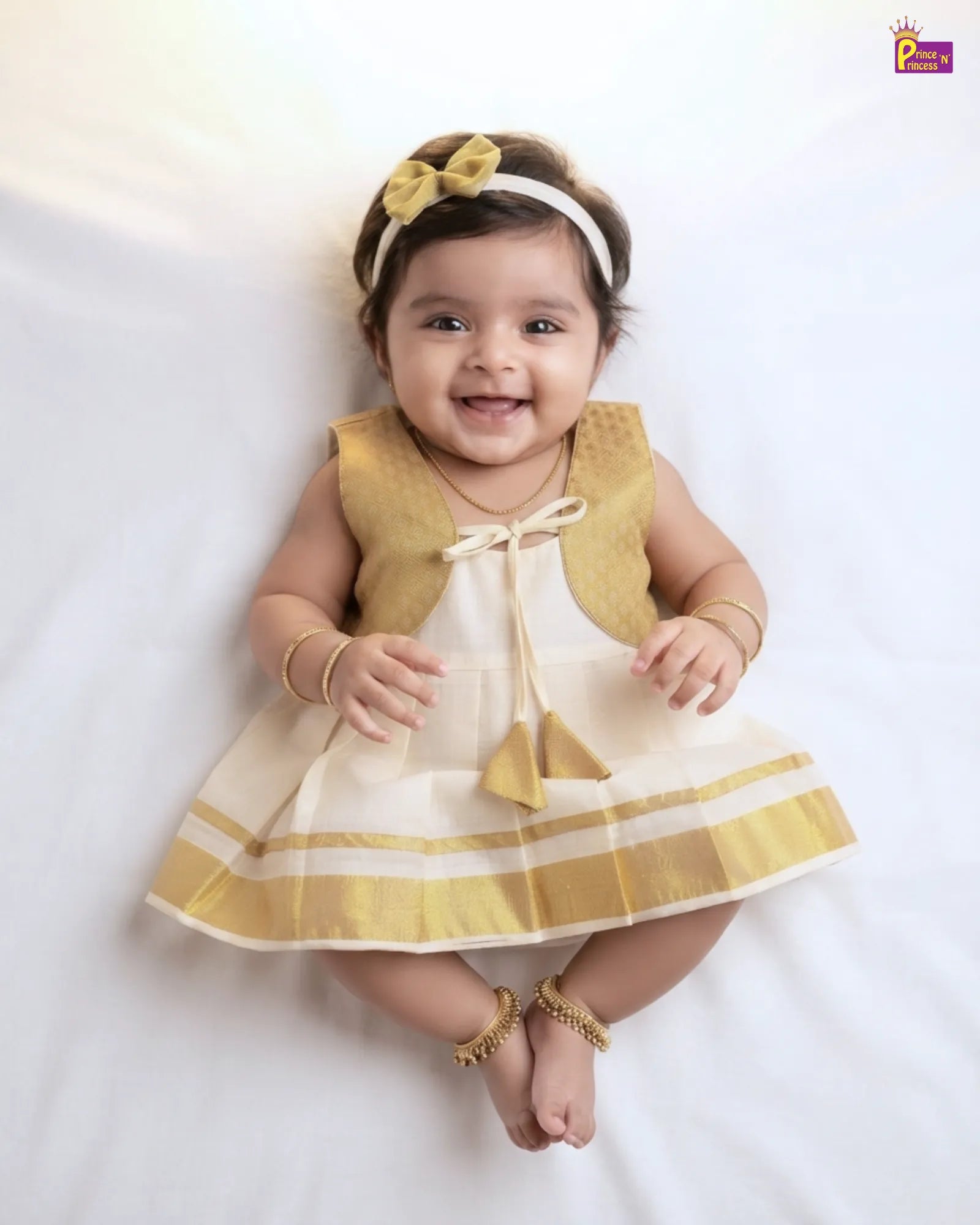 A smiling baby girl wearing a sleeveless cream and gold dress with a matching gold bow headband and gold jewelry, lying on a white fabric background.