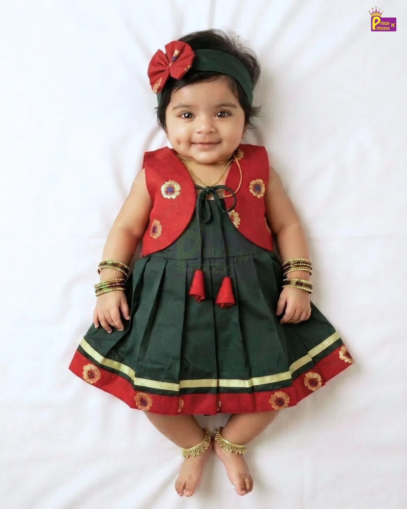 A baby girl wearing a traditional Indian ethnic dress costume, featuring a dark green pleated skirt with red and gold borders, a red sleeveless vest, and a matching dark green headband with a red bow. She is lying on a white sheet.