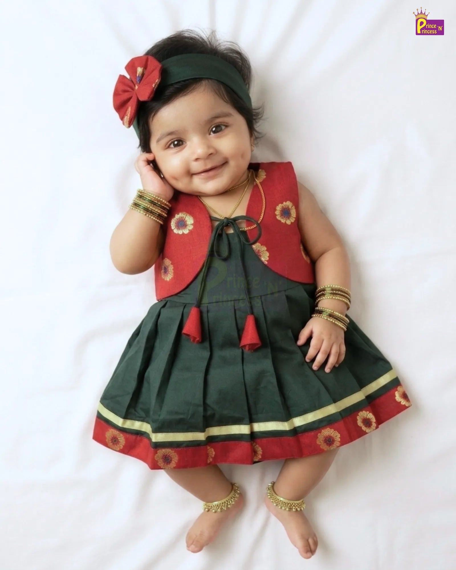 A baby girl lying on a white sheet wearing a dark green and red traditional Indian dress with gold floral embroidery, a red bow headband, and gold bangles and ankle bracelets.