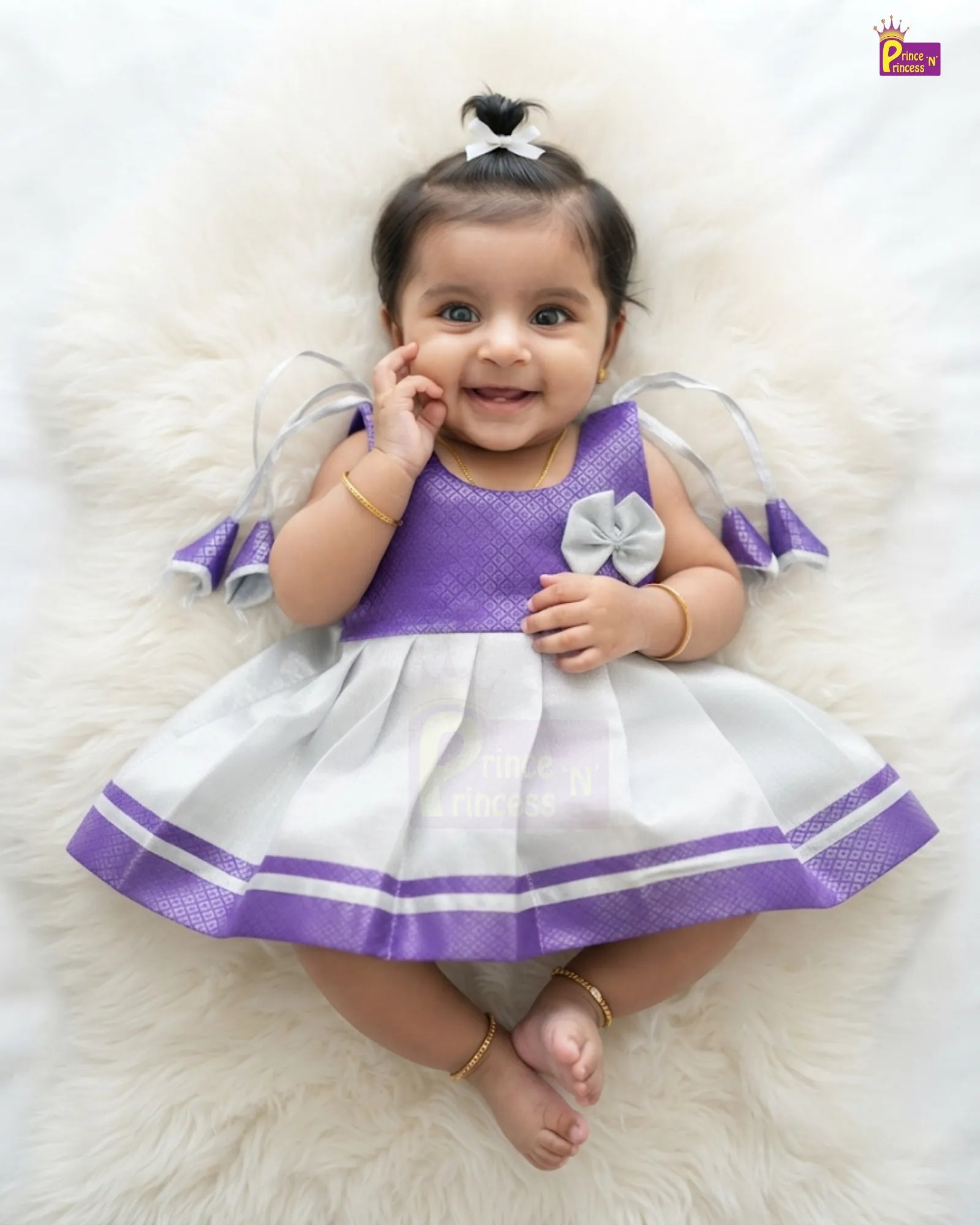 A baby girl lying on a white faux fur rug, wearing a purple and white dress with a white bow in her hair. The dress has a purple mesh bodice, a white pleated skirt with purple stripes, and greyish-white ruffled sleeves.